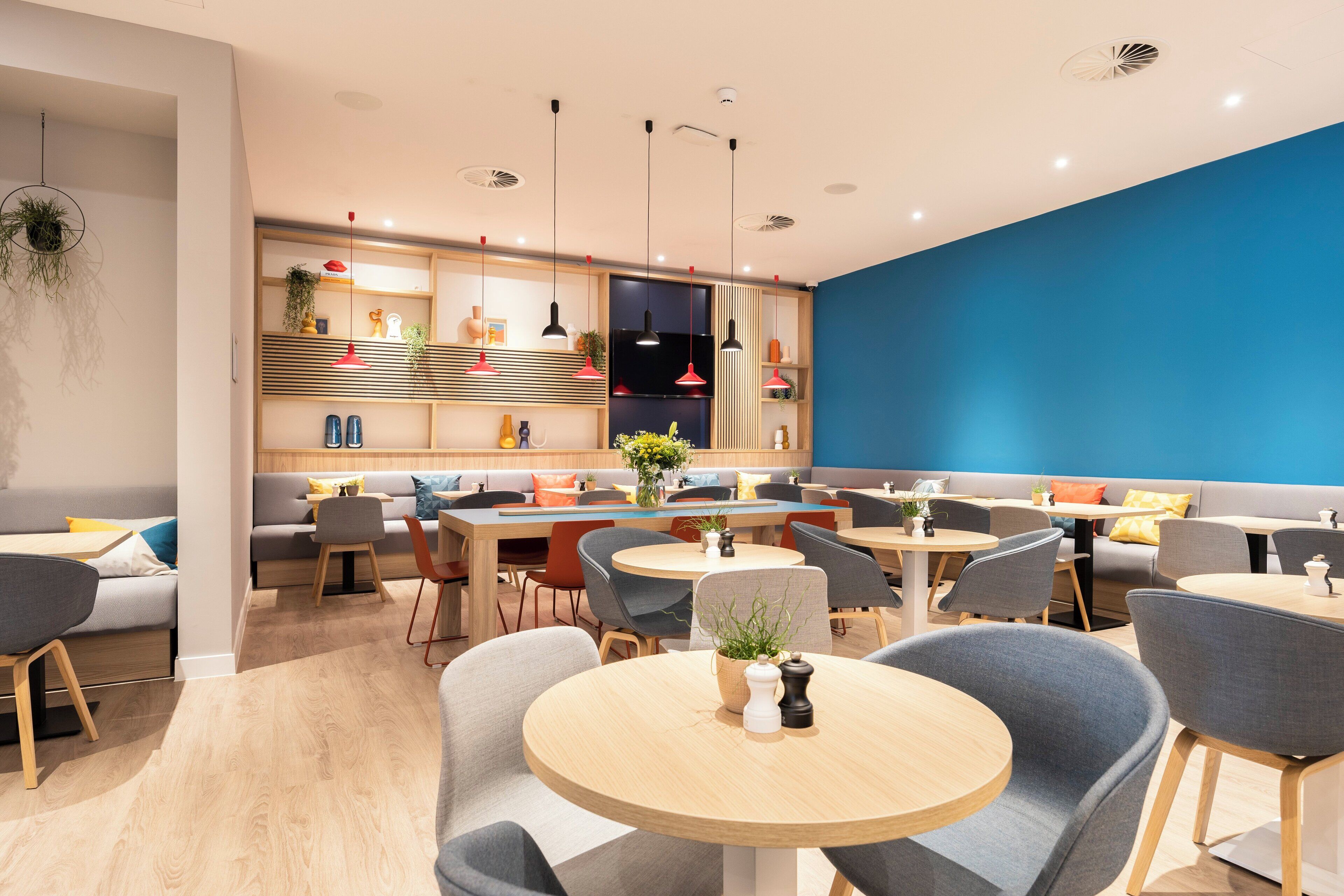 Un restaurante moderno con mesas redondas, sillas tapizadas en gris y una pared azul decorativa.