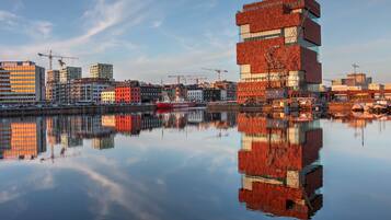 A waterfront cityscape with modern buildings, a red ship, and cranes, reflecting on the calm water.