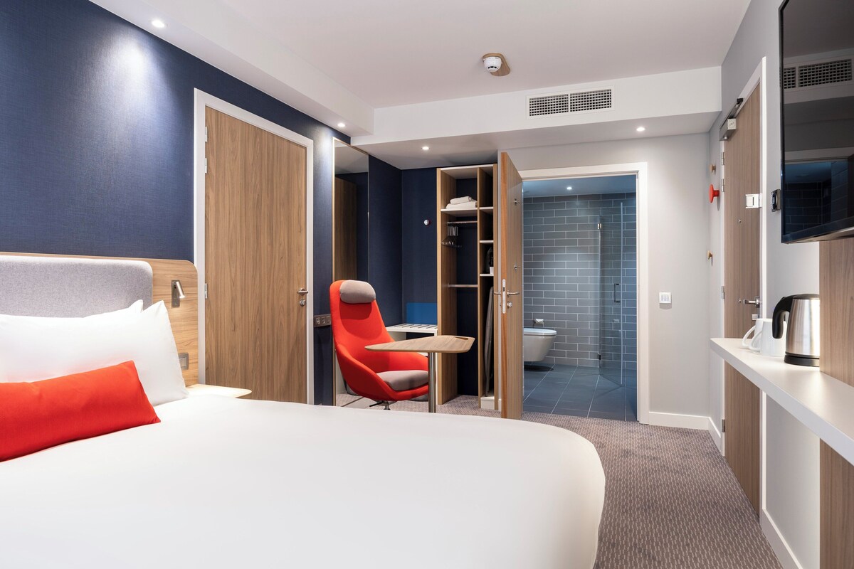 Habitación de hotel con cama, una silla roja, una mesita, armario y baño visible a través de una puerta abierta.