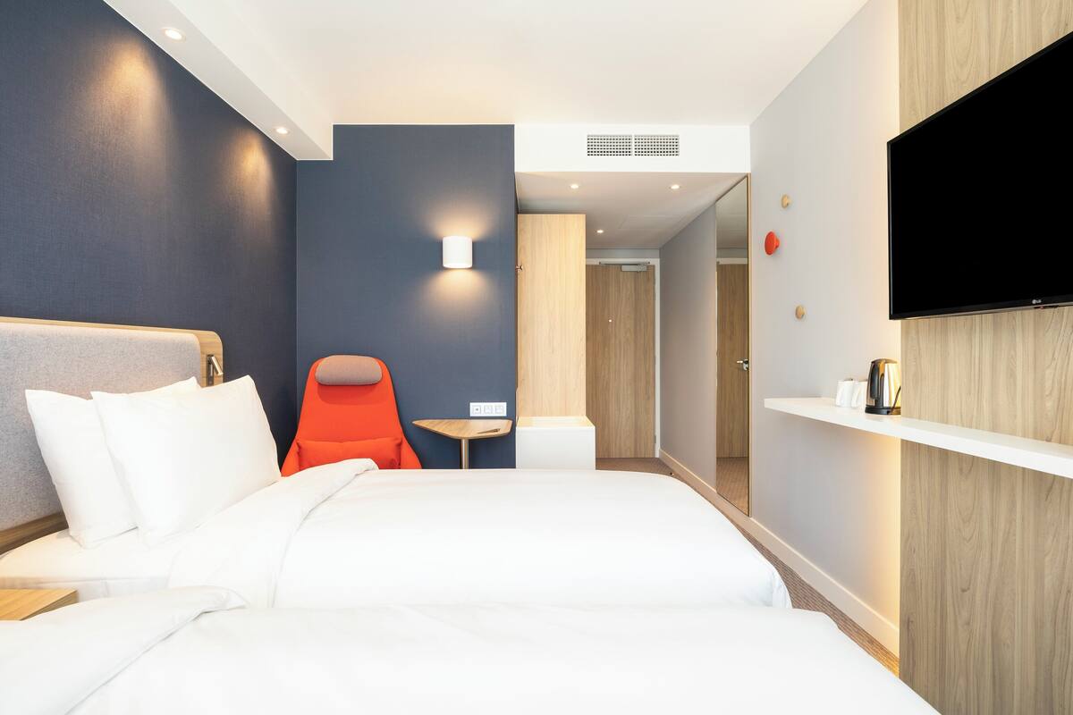A modern hotel room with a large bed, a small table, a chair, a TV mounted on the wall, and a wooden accent wall.