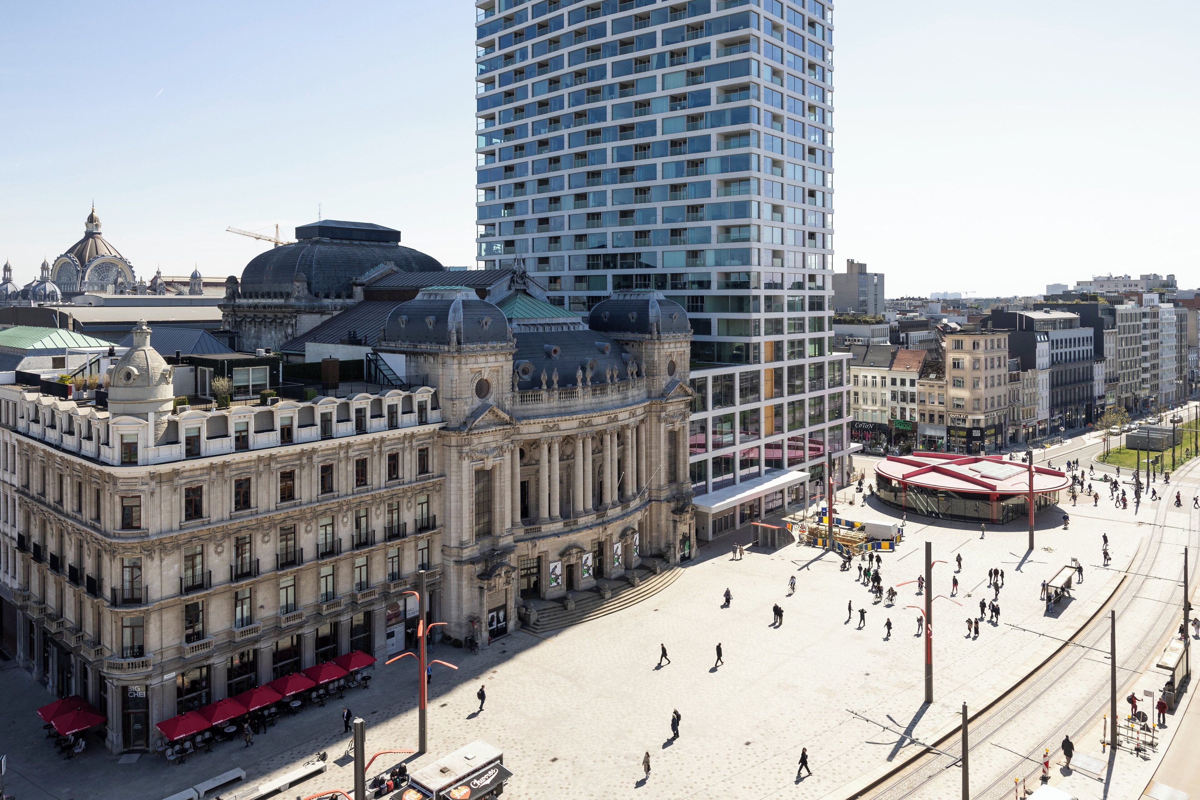 A historic building with classical architecture, a modern high-rise, and a public square with people.