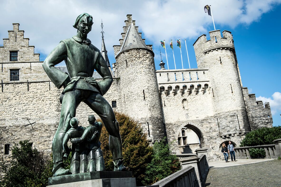 A statue of a knight in front of a castle with two towers and flags.