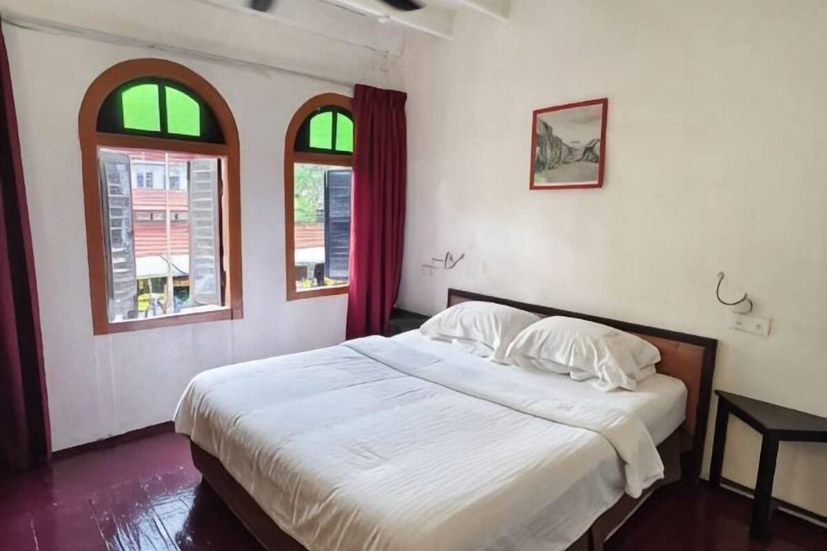 Una camera da letto con un letto, due finestre con tende, un quadro appeso e un tavolino.