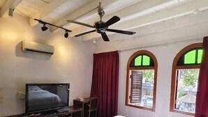A hotel room with a bed, a desk with a television, a ceiling fan, and two windows with curtains.