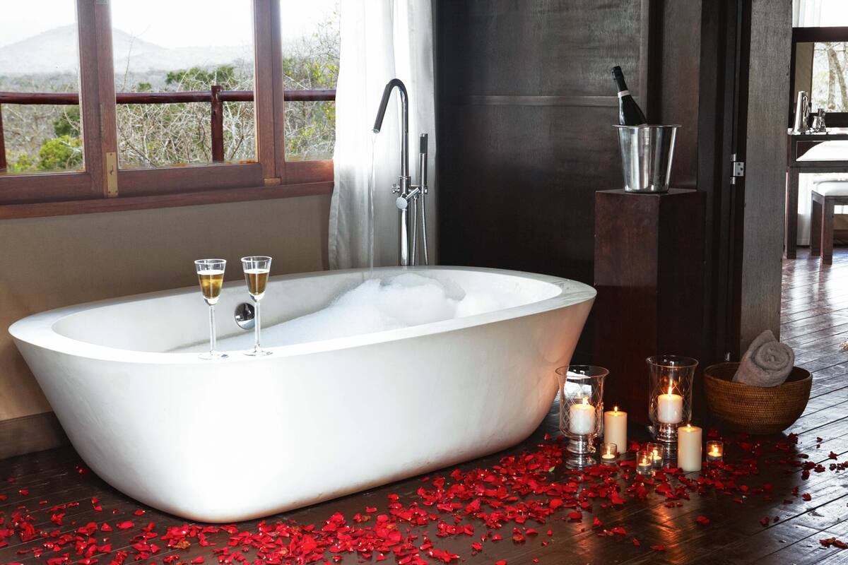 A large freestanding bathtub with champagne glasses, candles, and scattered rose petals.
