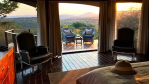 A room with a bed, two chairs, a small table with bottles, and a view of the landscape. - Thanda Safari (Hluhluwe)