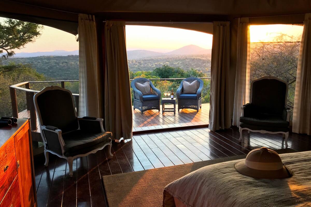 A room with a bed, two chairs, a small table with bottles, and a view of the landscape.