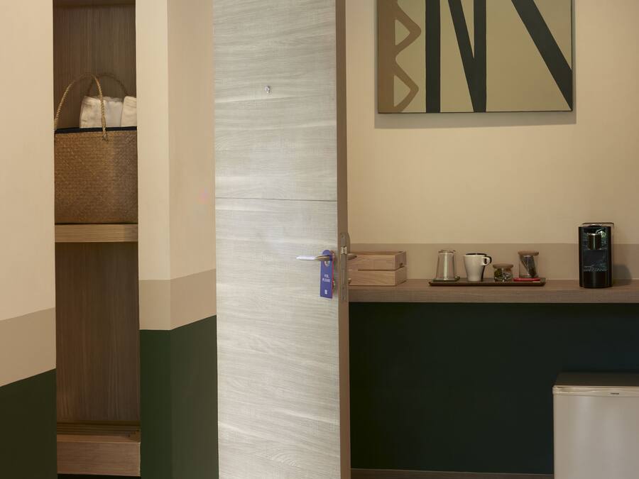 A hotel room with a wooden wardrobe, a kitchenette with a coffee machine, and a wall-mounted artwork.