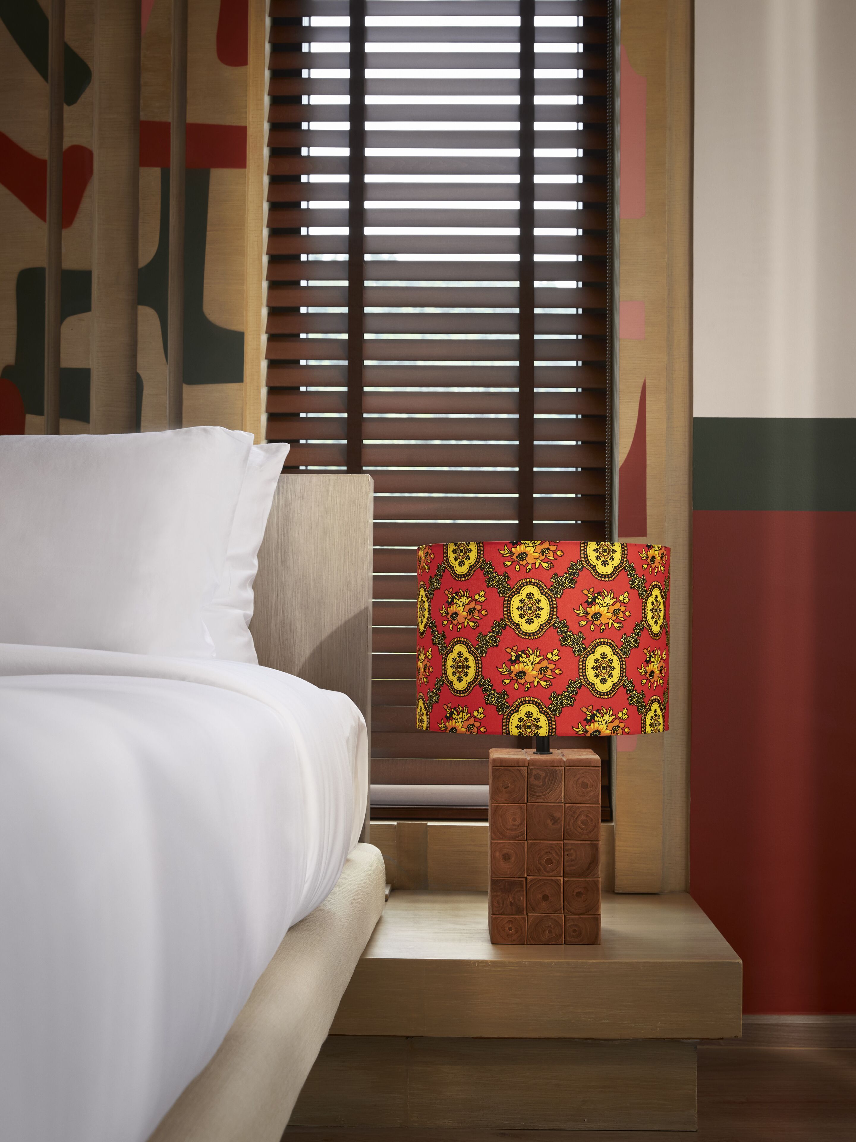 A neatly made bed with white linens, a wooden nightstand with a patterned tablecloth, and a window with blinds.
