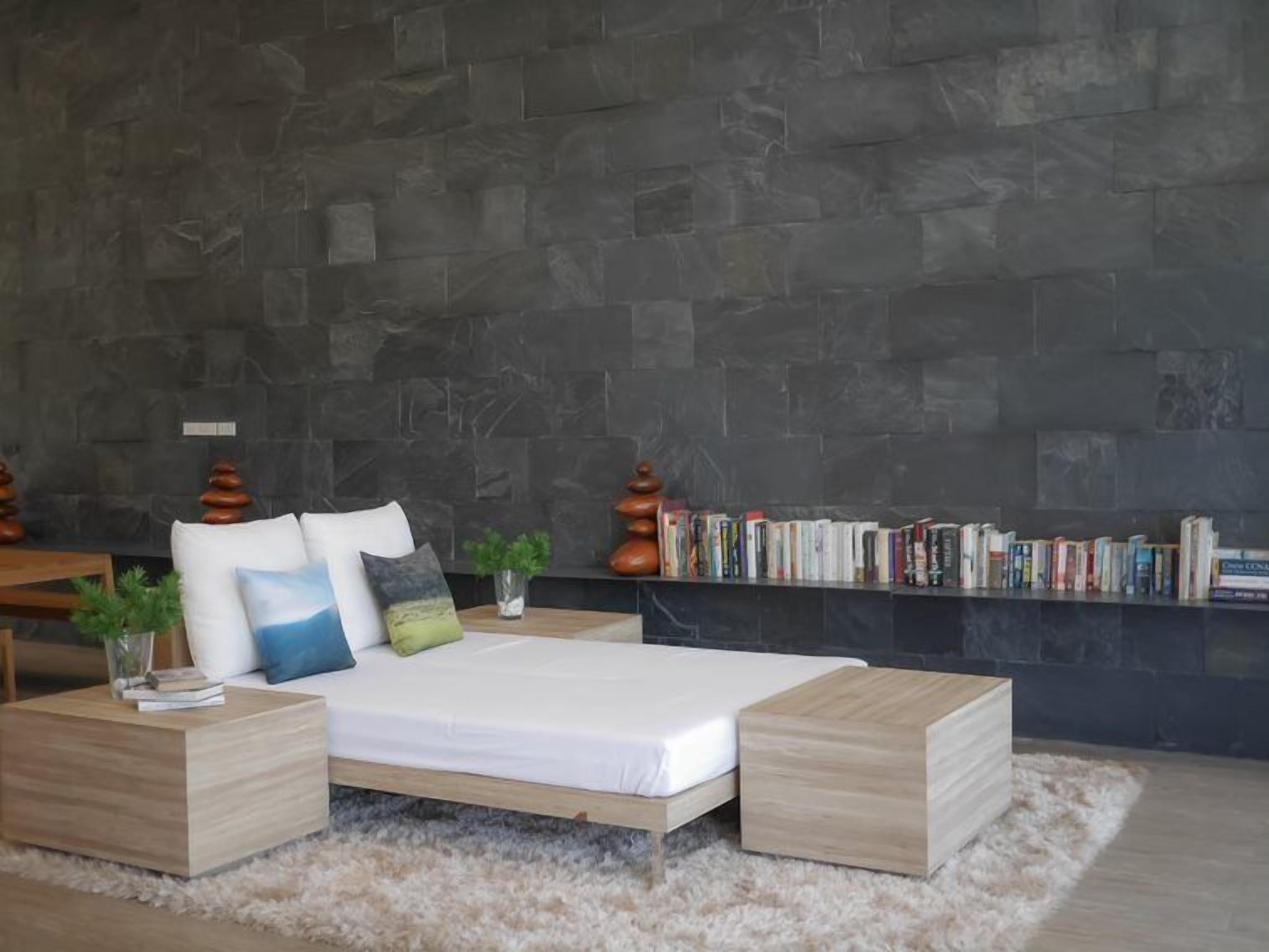 Un dormitorio moderno con pared de piedra, estructura de cama de madera, colchón blanco y estantería llena de libros.