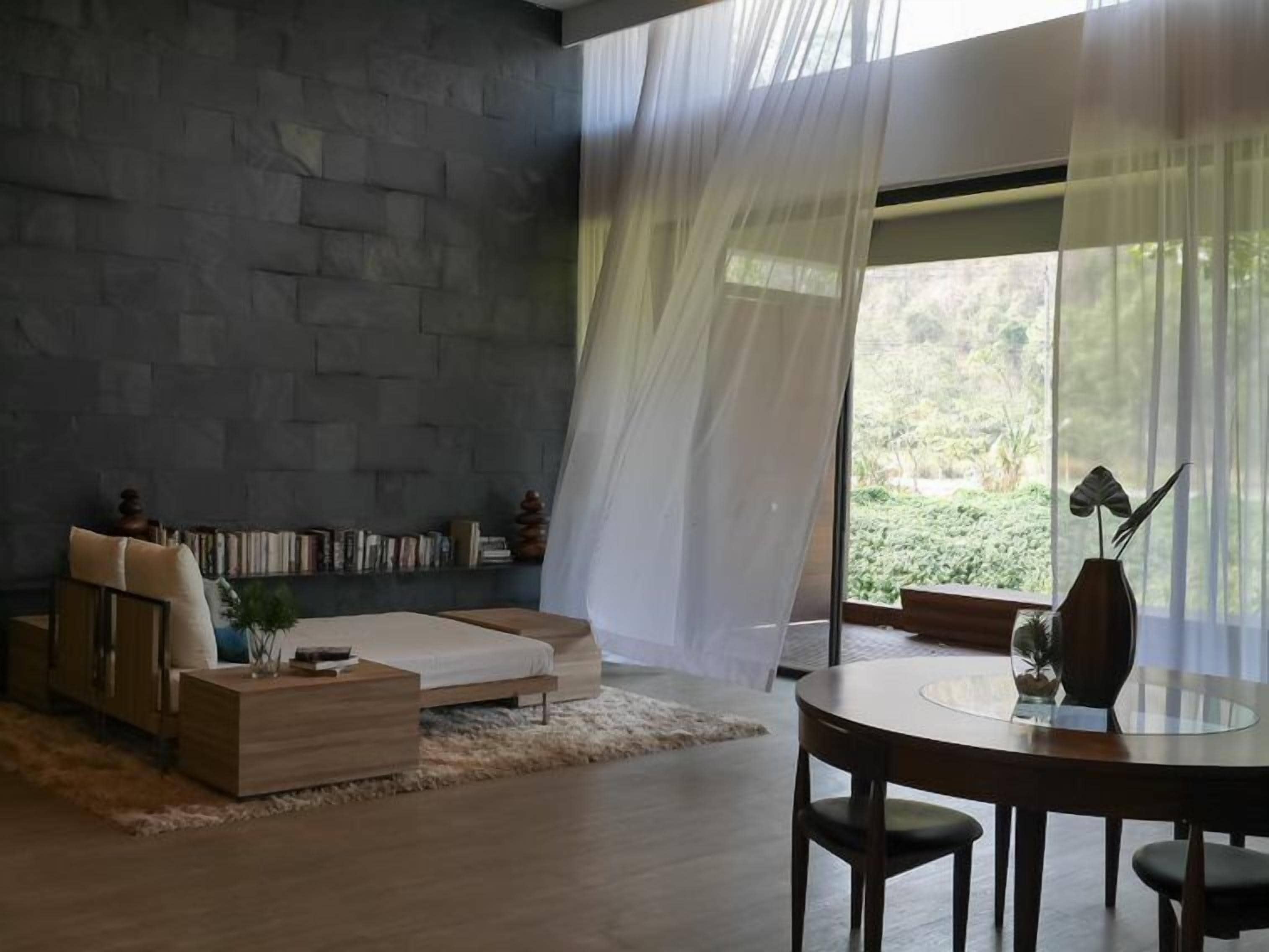 A modern living room with a stone wall, a sofa, a coffee table, a dining table with chairs, and sheer curtains.