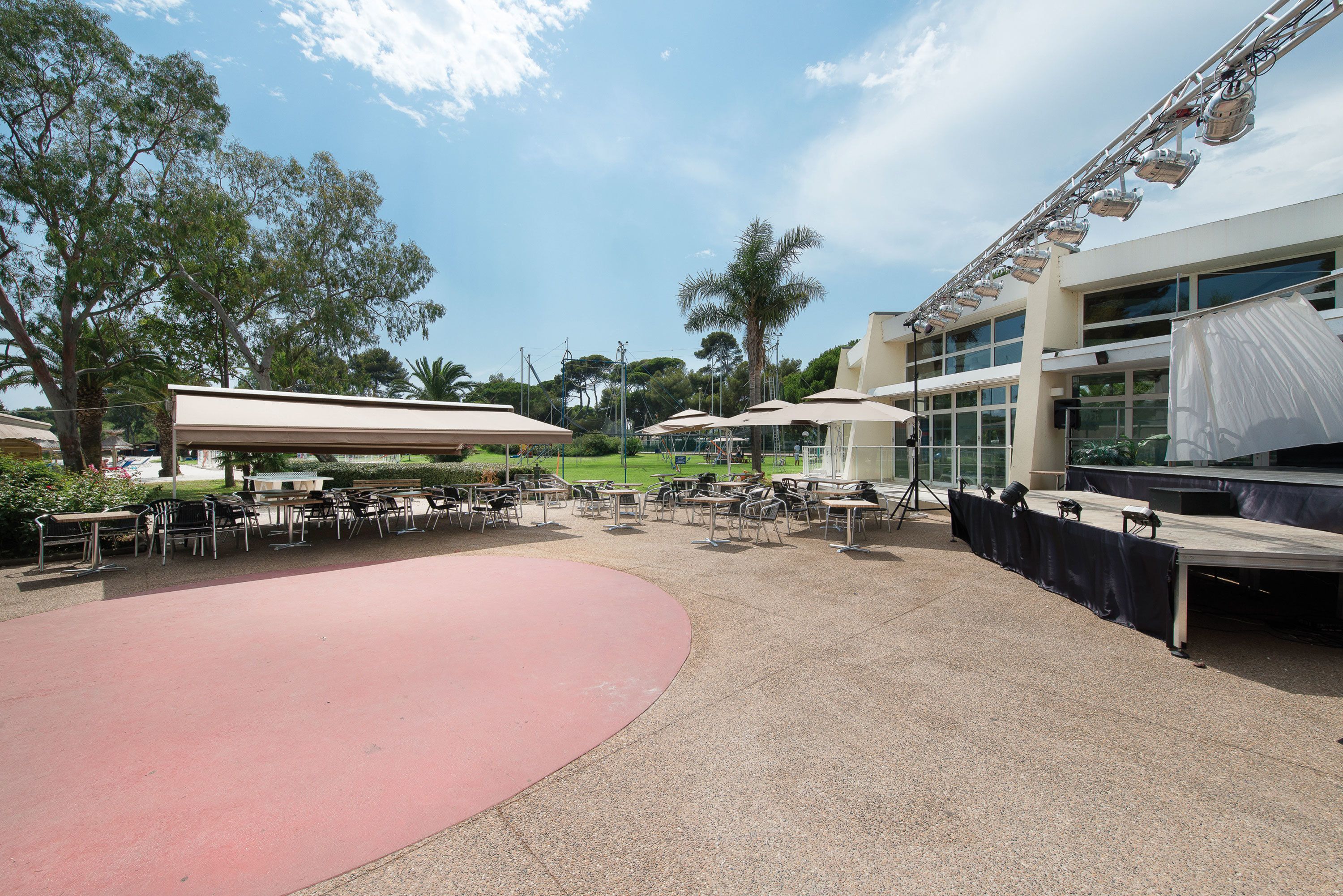 An outdoor patio area with tables and chairs, a stage with lighting equipment, and a building with large windows in the background.