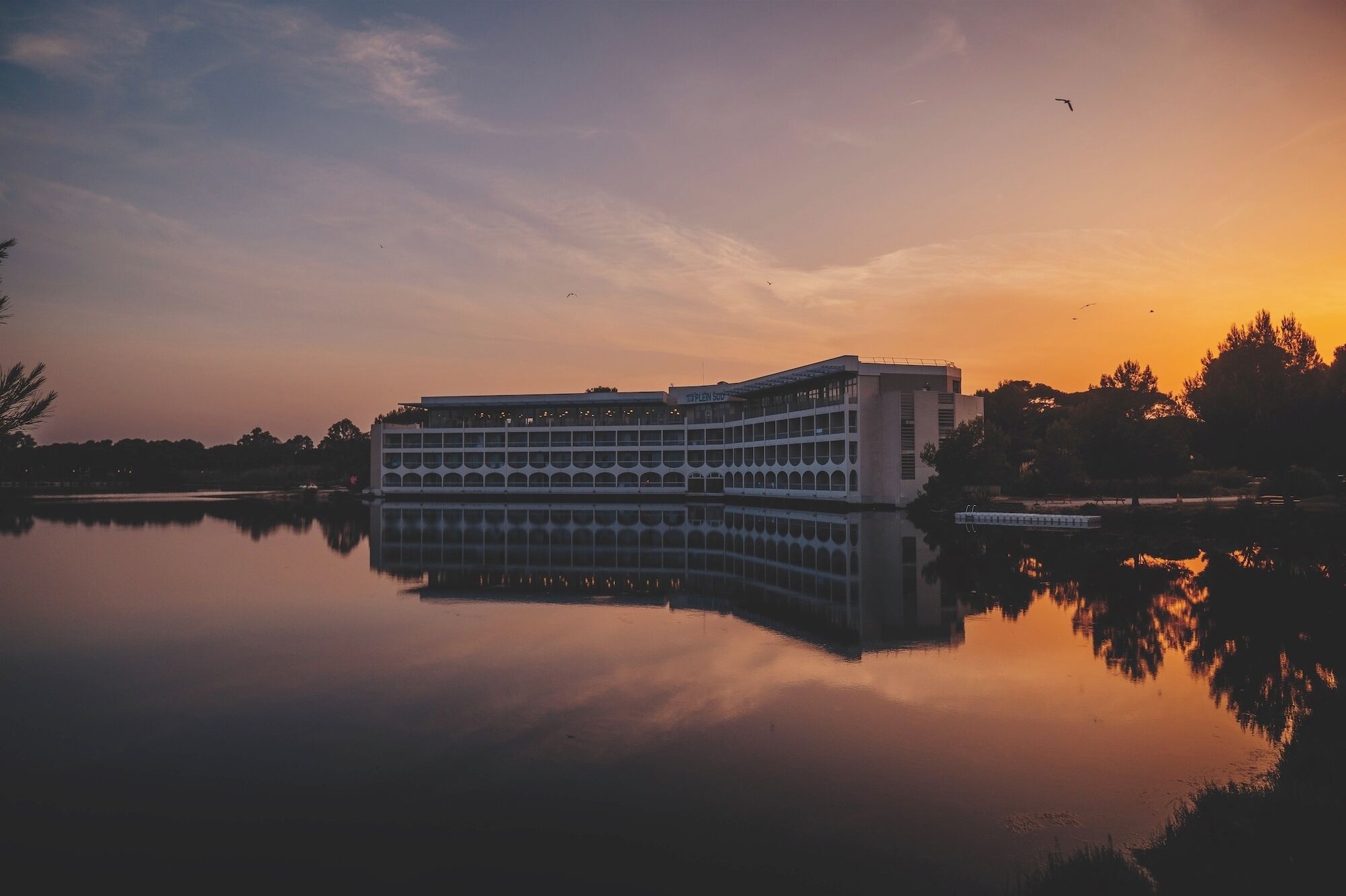 Un grand hôtel à plusieurs étages, dont l’image se reflète dans l’eau, entouré d’arbres et illuminé par un ciel au coucher du soleil.