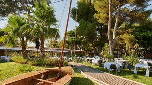 A boat docked near a wooden deck, surrounded by tables and chairs set up for dining, with palm trees and a building in the background. - Club Vacances Bleues Plein Sud (Hyères)