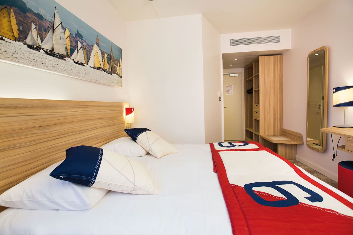 A hotel room with a bed, pillows, a painting, a wardrobe, and a mirror.