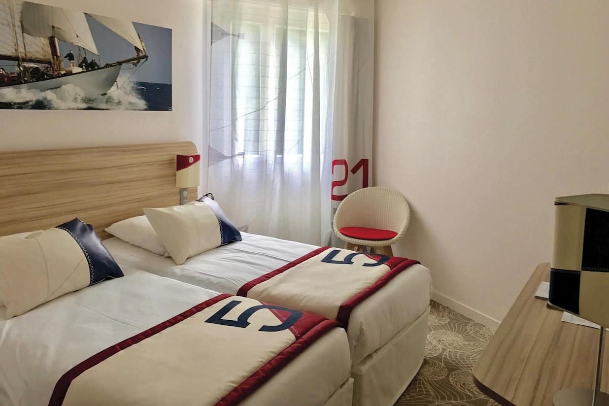 A hotel room with two beds, a wooden headboard, a window with sheer curtains, a wicker chair, and a small table.