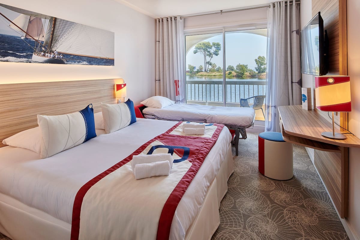 A hotel room with two beds, a TV, a desk, and a balcony with a view of the sea.