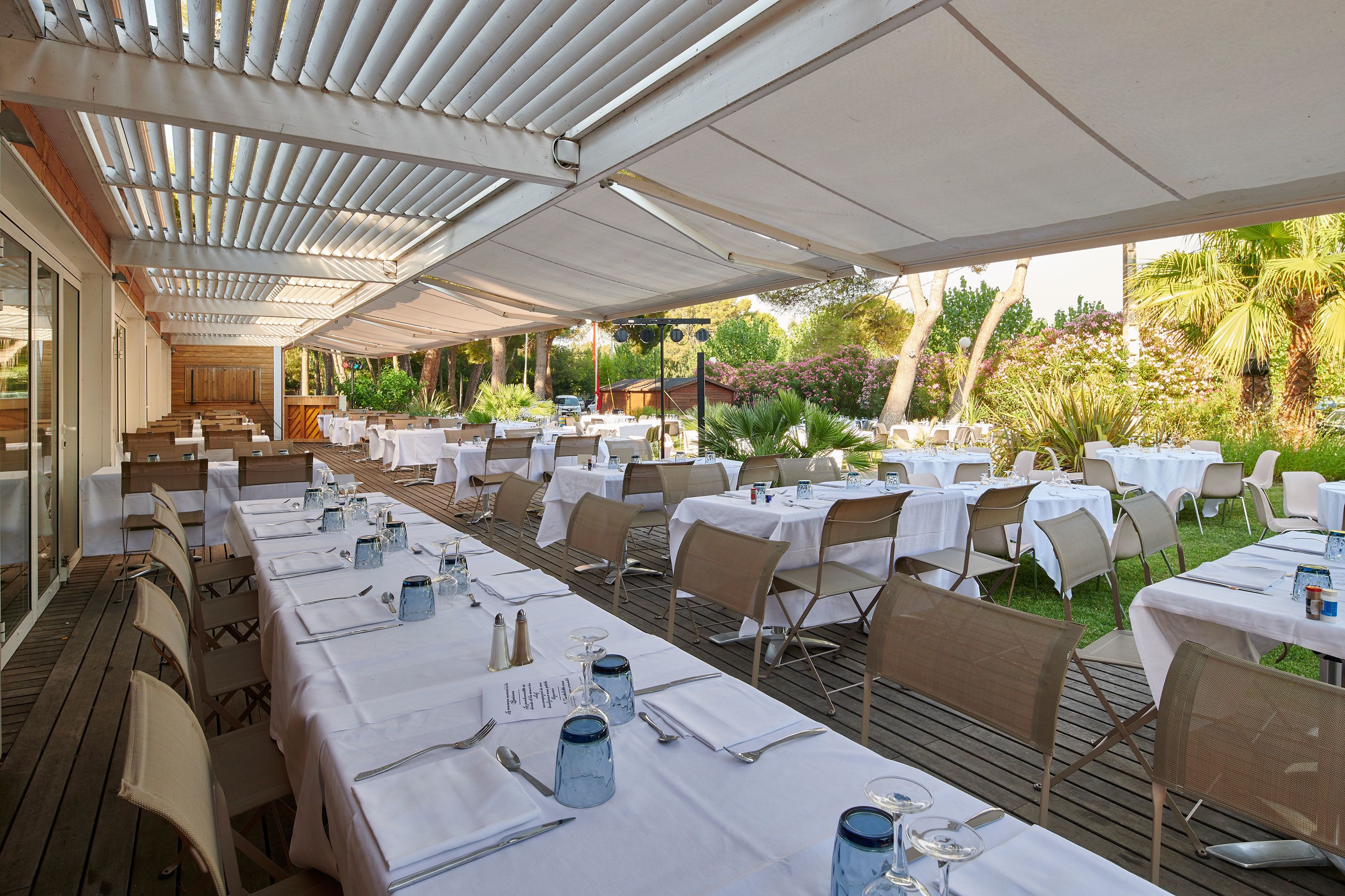 An outdoor dining area with tables set for a meal, surrounded by greenery and a covered walkway.