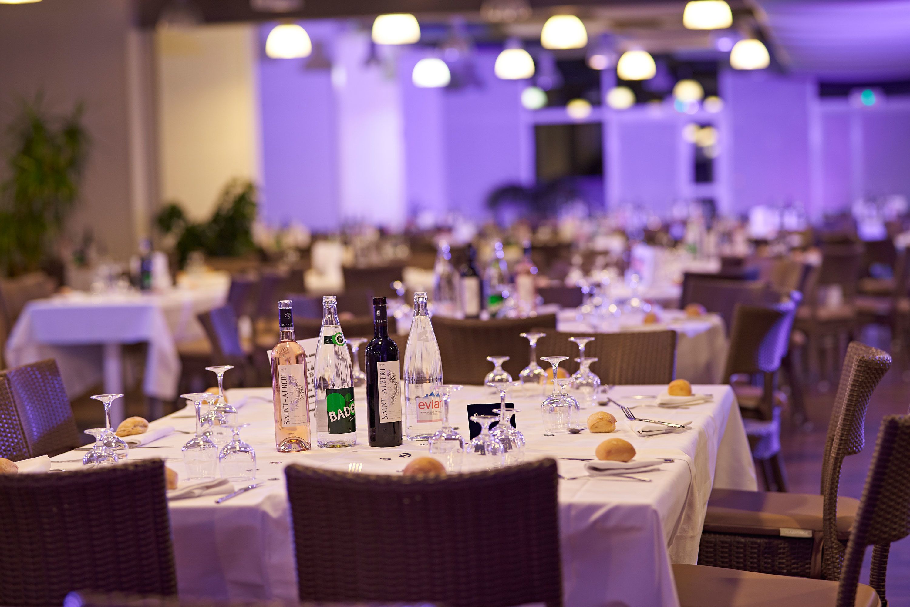 A banquet hall with multiple tables set for dining, featuring wine bottles and glasses.