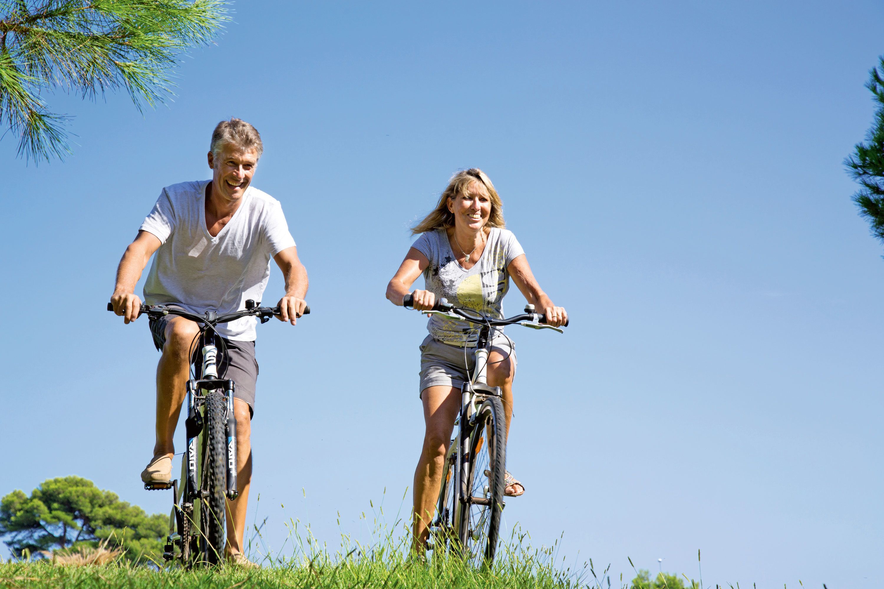 Two people riding bicycles on a grassy field with clear skies.
