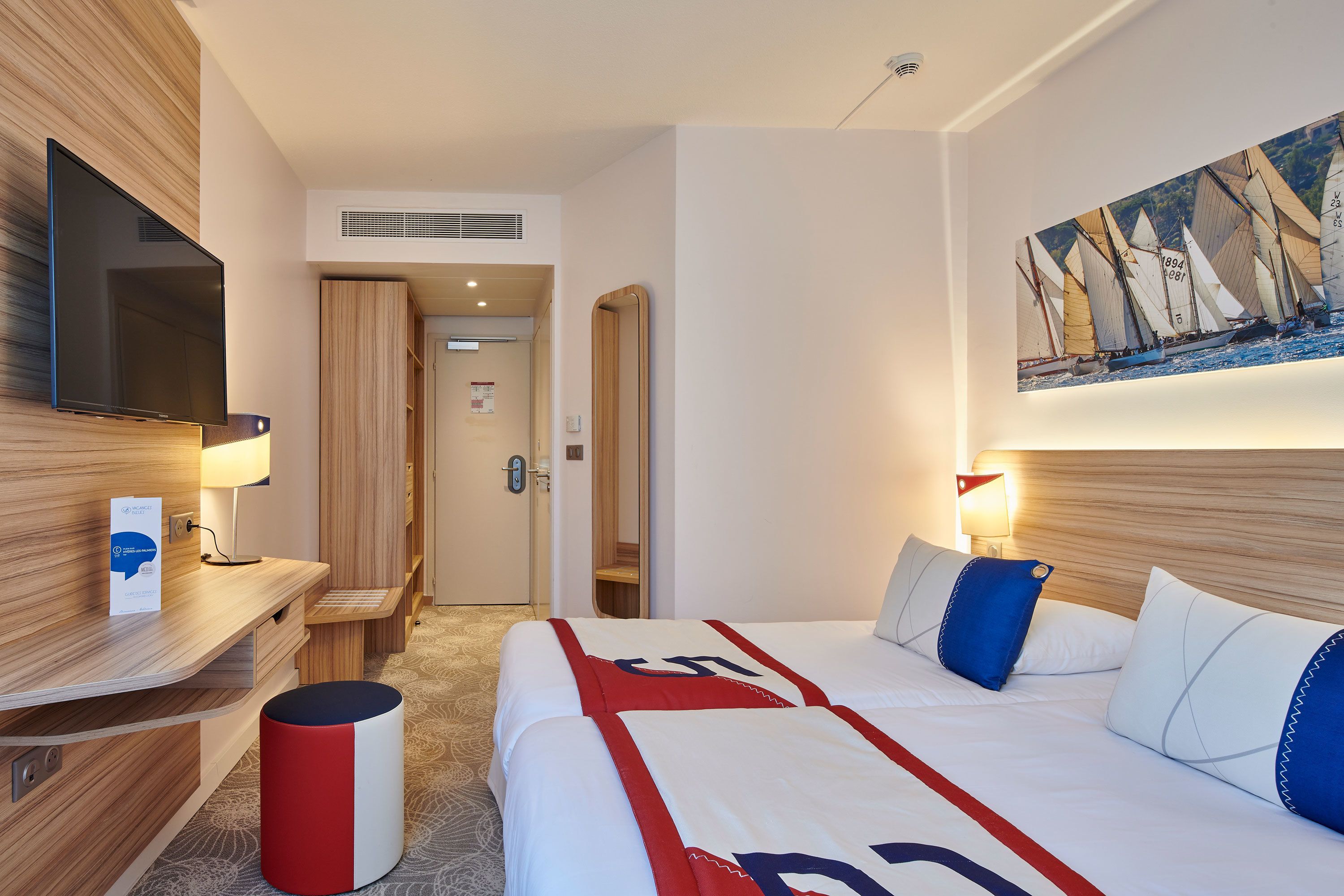 A hotel room with a bed, bedside table, TV, and a painting of sailboats.