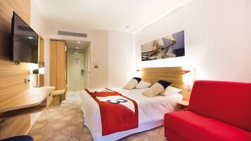 A hotel room with a bed, a red sofa, a TV mounted on the wall, and a painting above the bed.