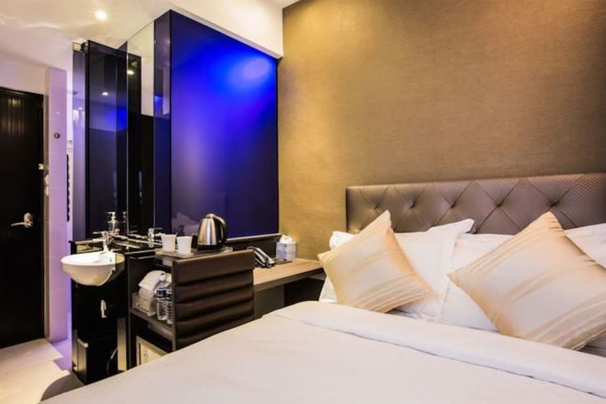 A modern hotel room with a large bed, a bedside table with a lamp, a mirror, and a bathroom with a sink and toilet.