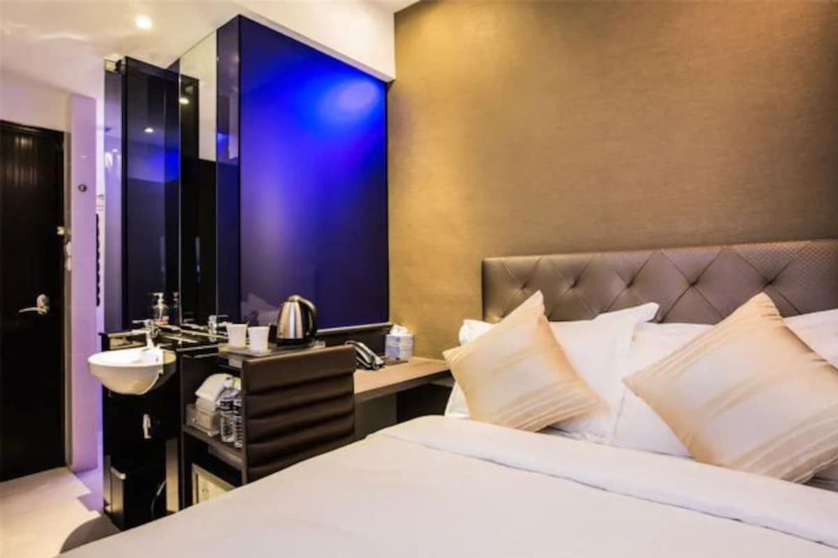 A modern hotel room with a large bed, a bedside table, a built-in vanity with a mirror, and a wall-mounted TV.