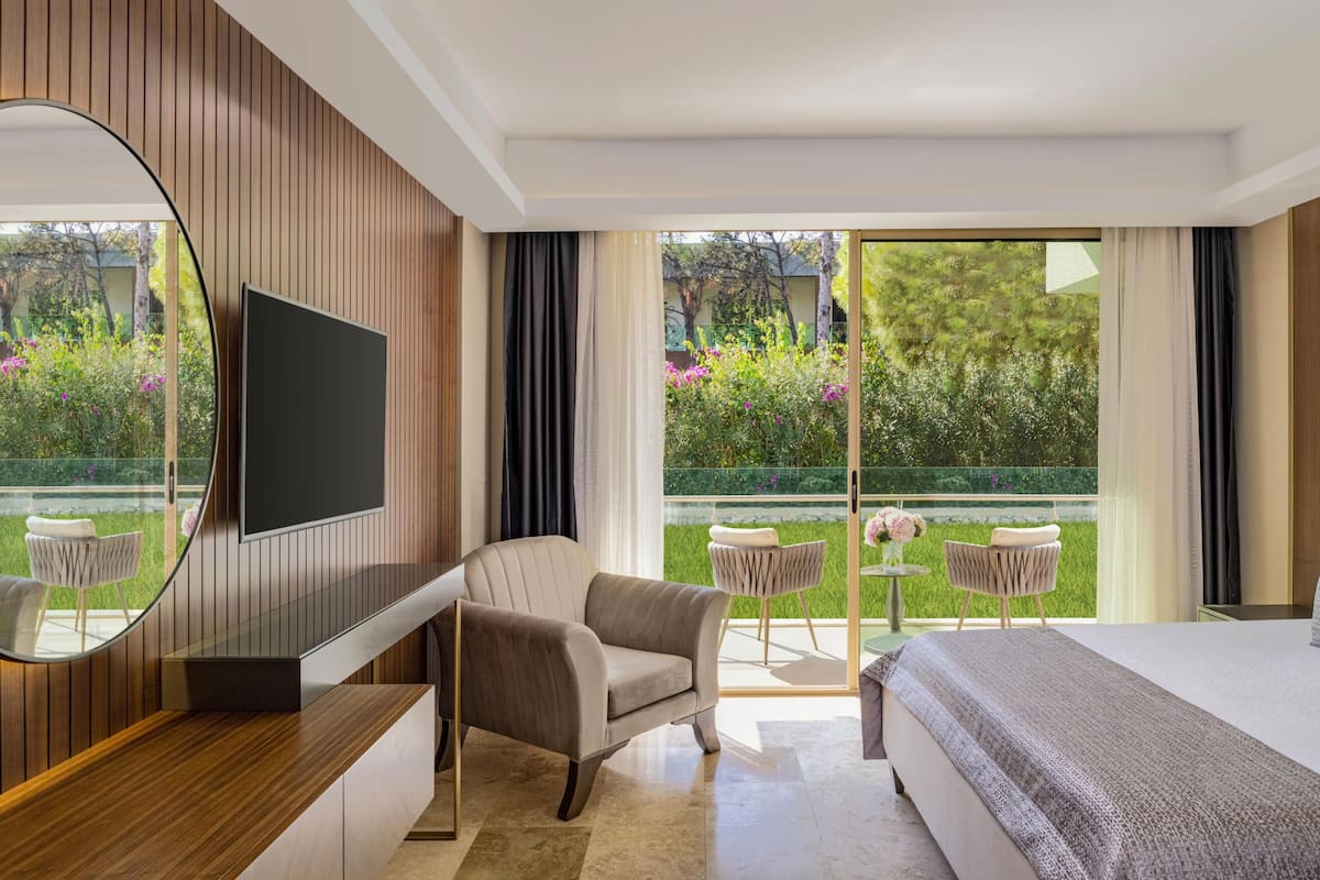 Una habitación de hotel moderna con una cama grande, televisión, espejo redondo y balcón con mobiliario exterior.