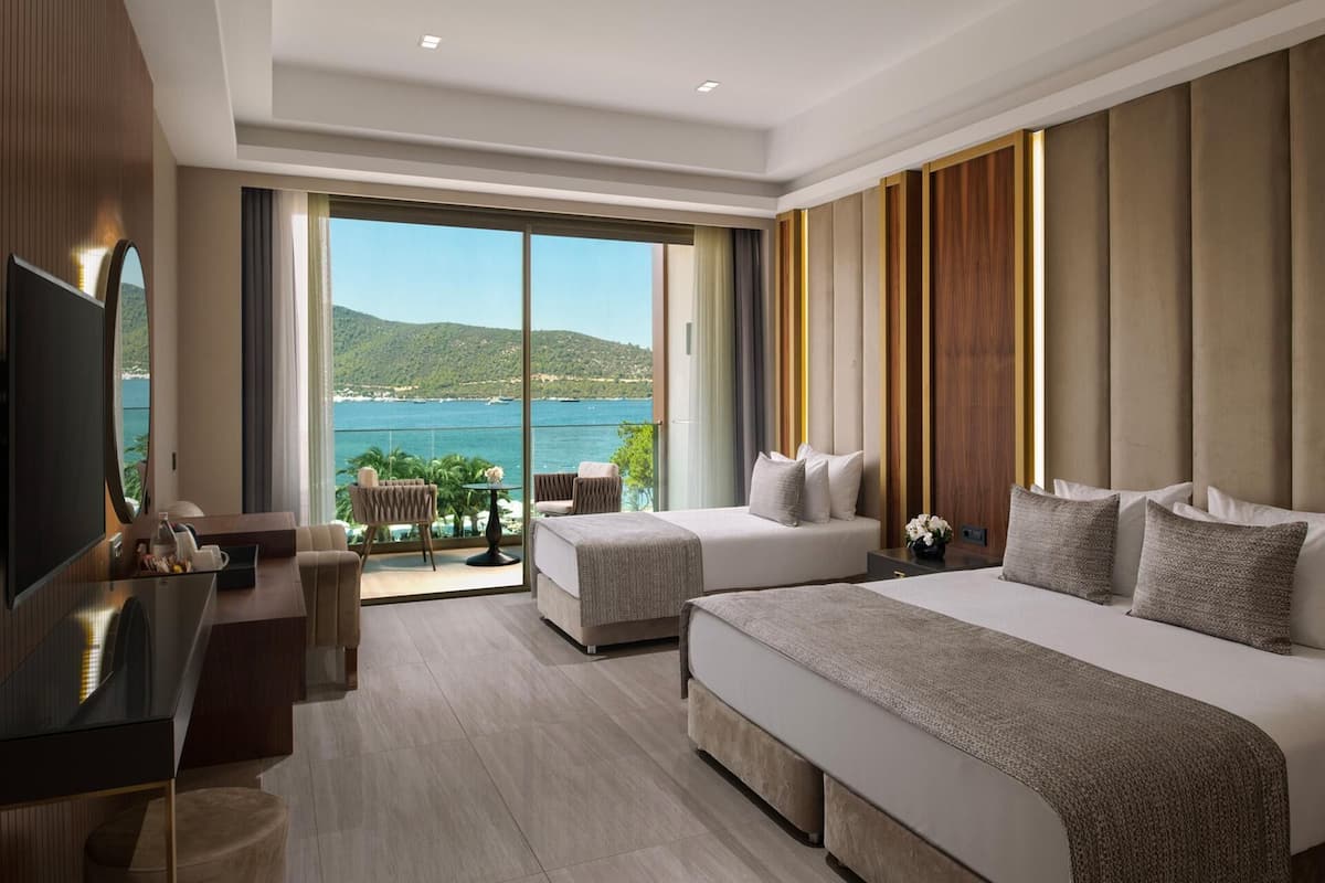 Habitación de hotel con dos camas, televisión, escritorio y balcón con vistas al mar.
