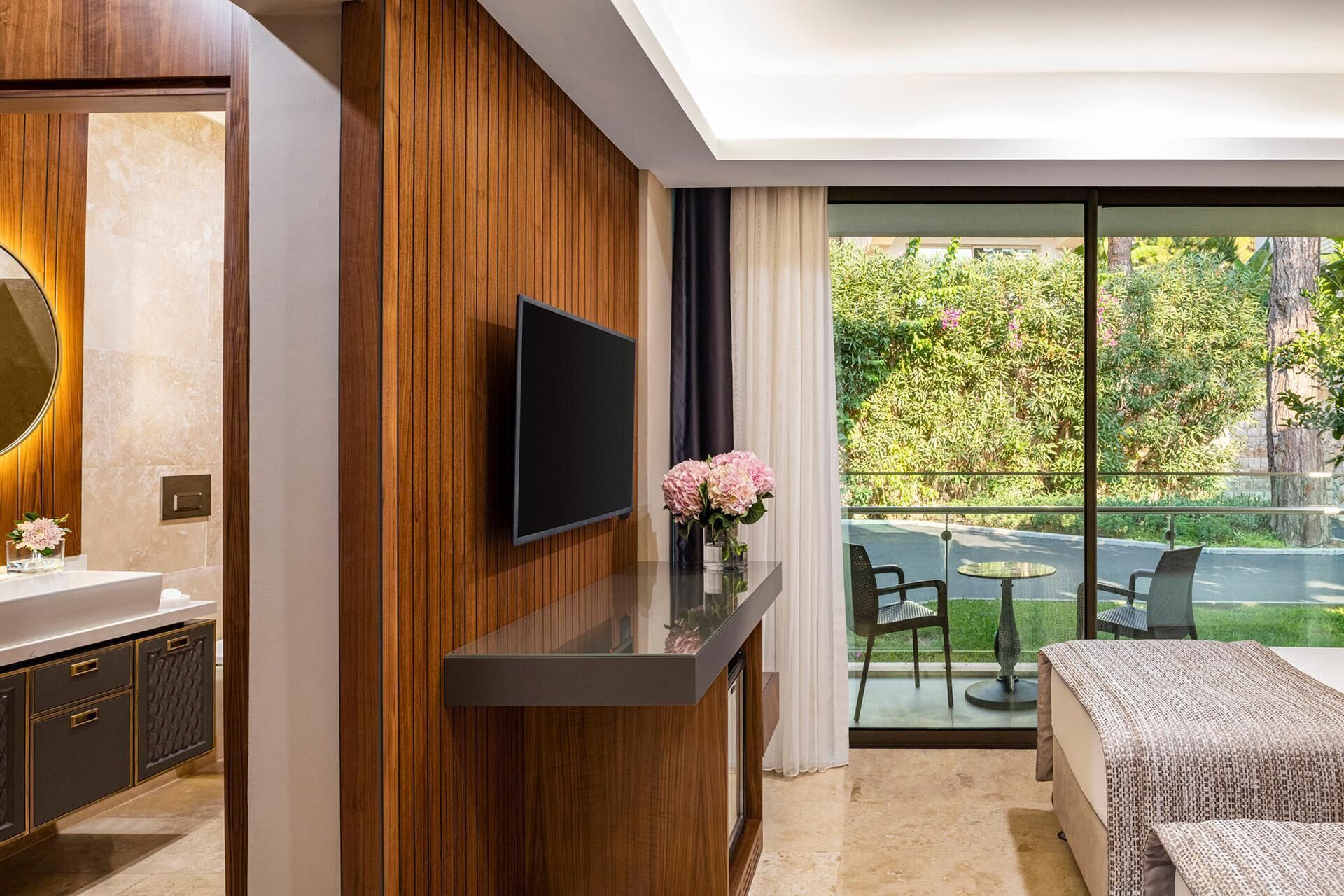 Una camera moderna con TV fissata a una parete rivestita in legno, un piccolo tavolo con sedie e un balcone affacciato sul verde.