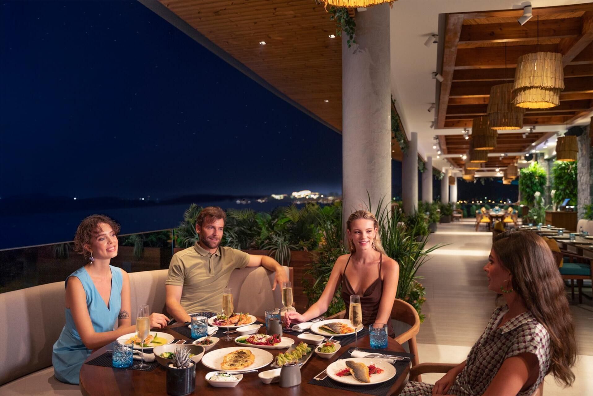 A dining area with a view of the ocean, featuring a table set with various dishes and drinks, surrounded by plants and wooden beams.
