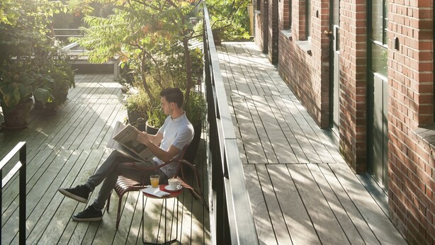 Une personne assise sur un banc en bois sur un balcon, avec un bâtiment en briques et de la verdure en arrière-plan.