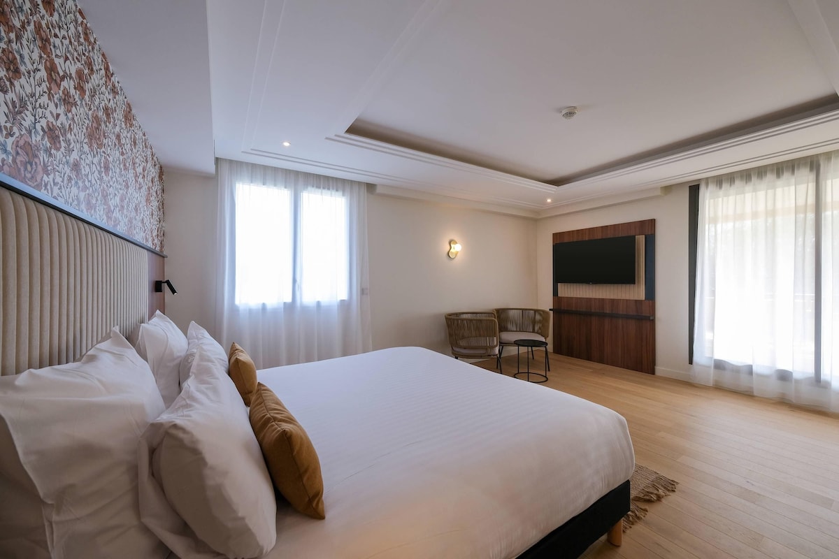 A hotel room with a large bed, a television, a seating area with chairs, and a window with curtains.