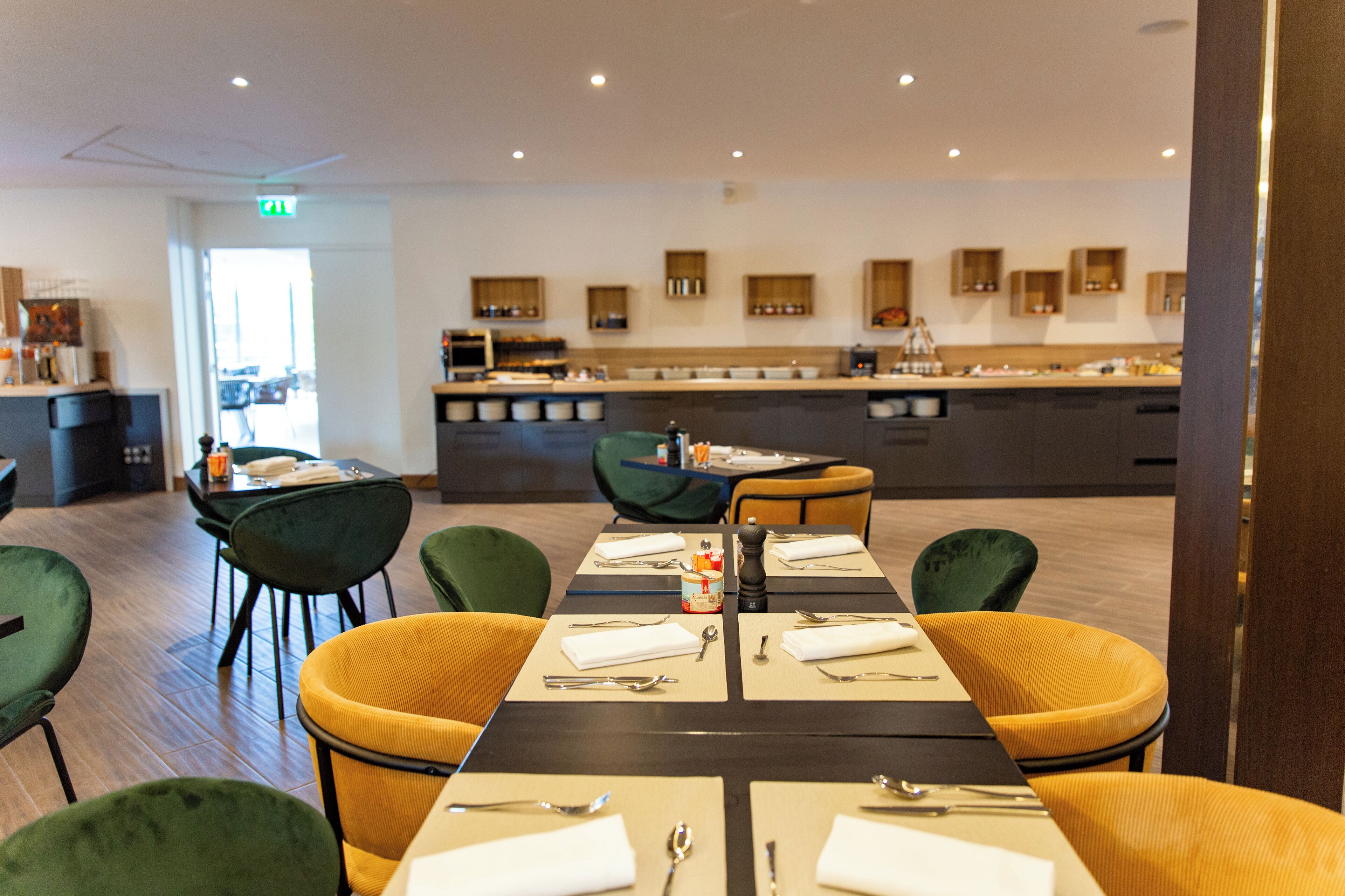 A modern dining area with a long table set for four, surrounded by green and yellow chairs.