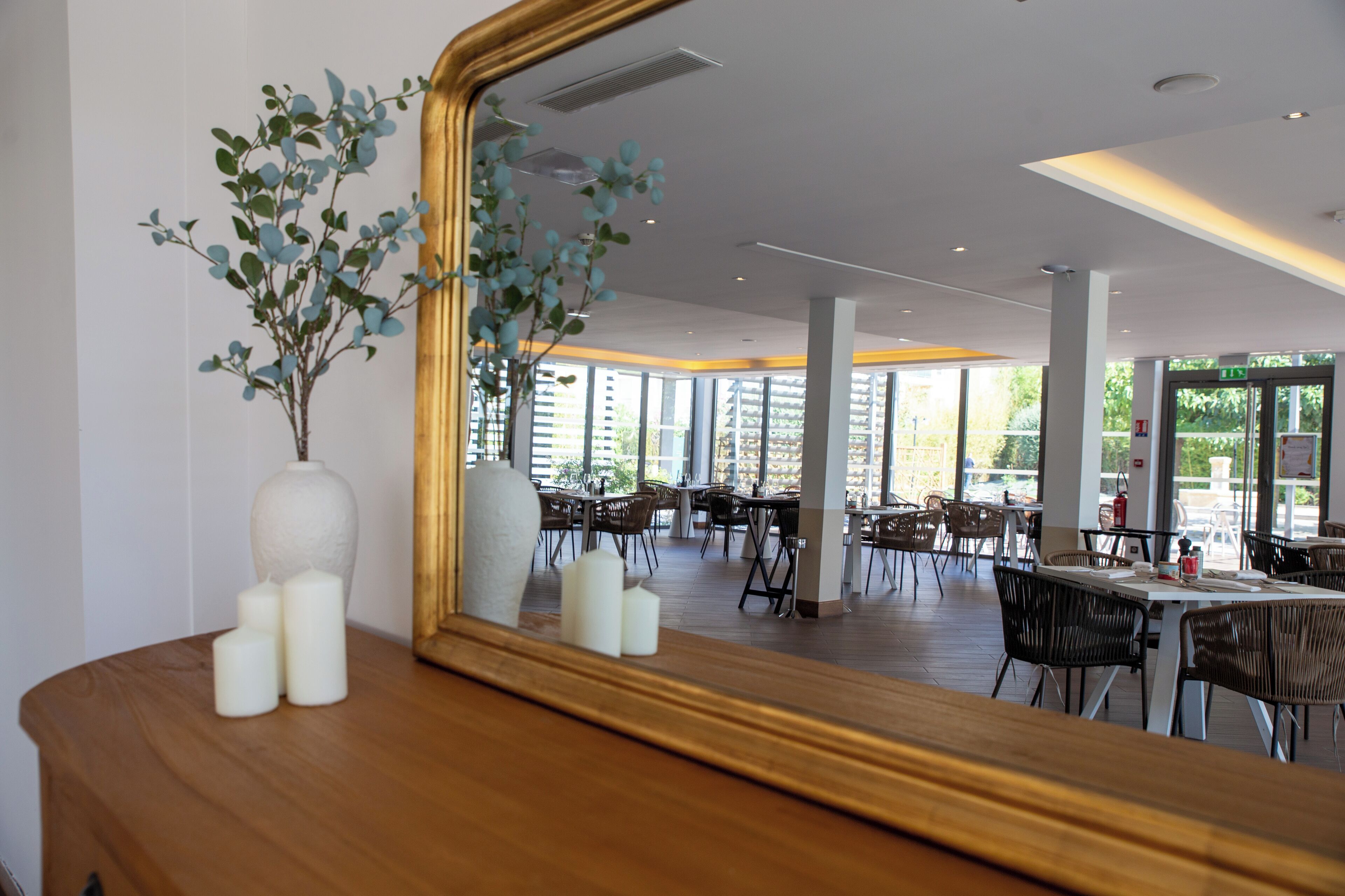 A modern dining area with wooden tables and chairs, a large mirror reflecting the space, and a vase with greenery on a wooden surface.
