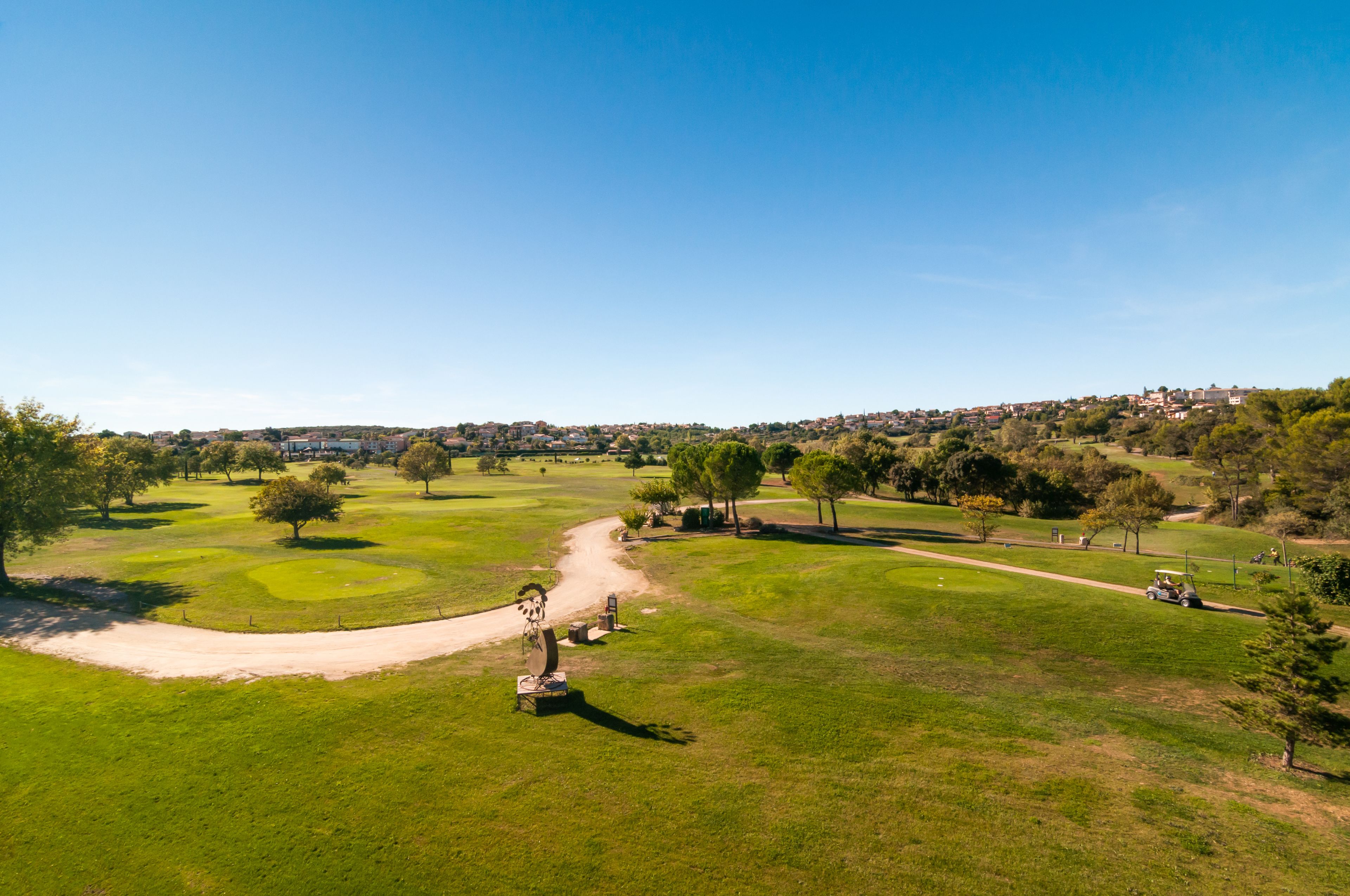 Un parcours de golf avec un tracé bien défini, des arbres et des bâtiments visibles au loin.
