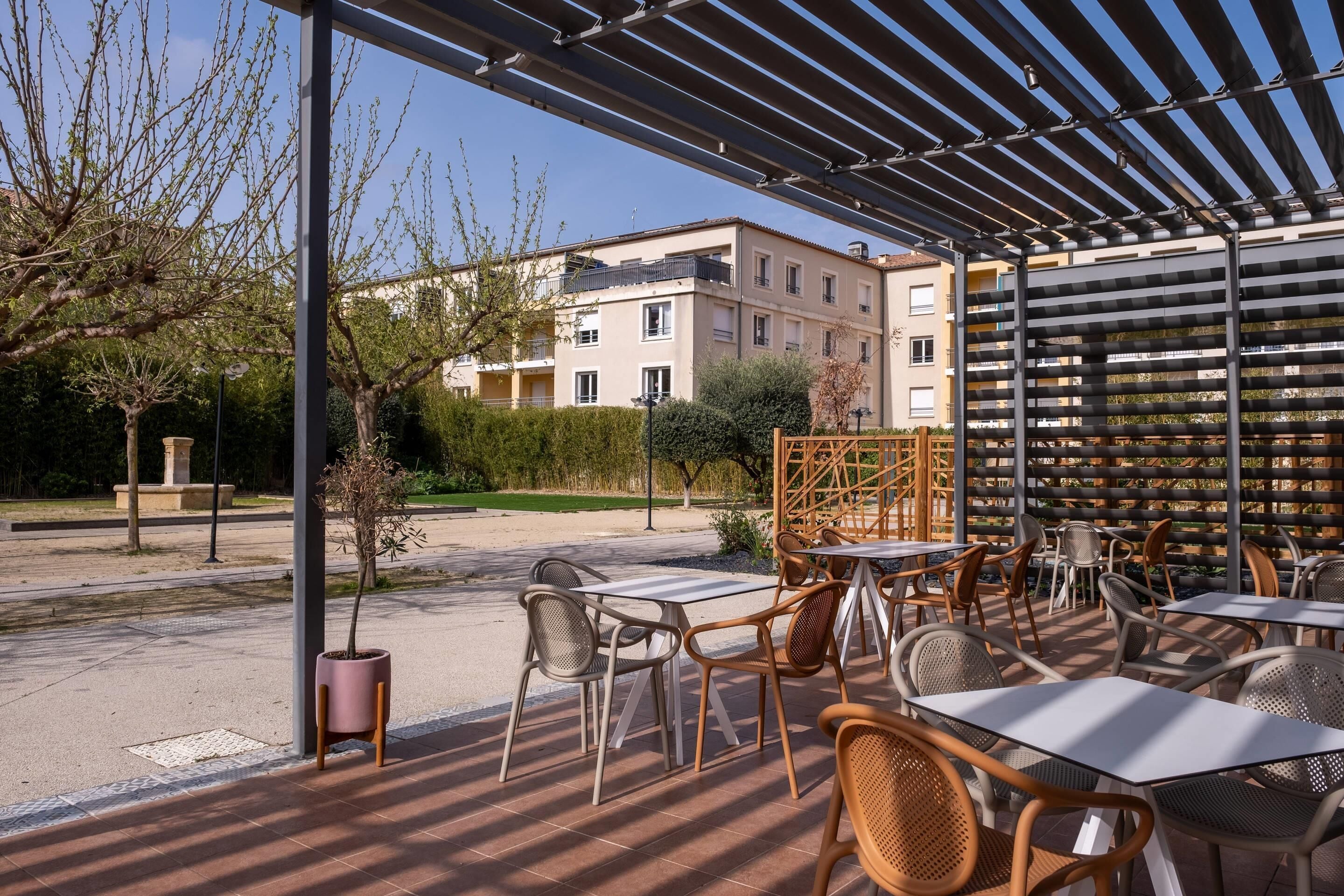 Un patio al aire libre con mesas y sillas, una pérgola y vistas de un edificio residencial al fondo.