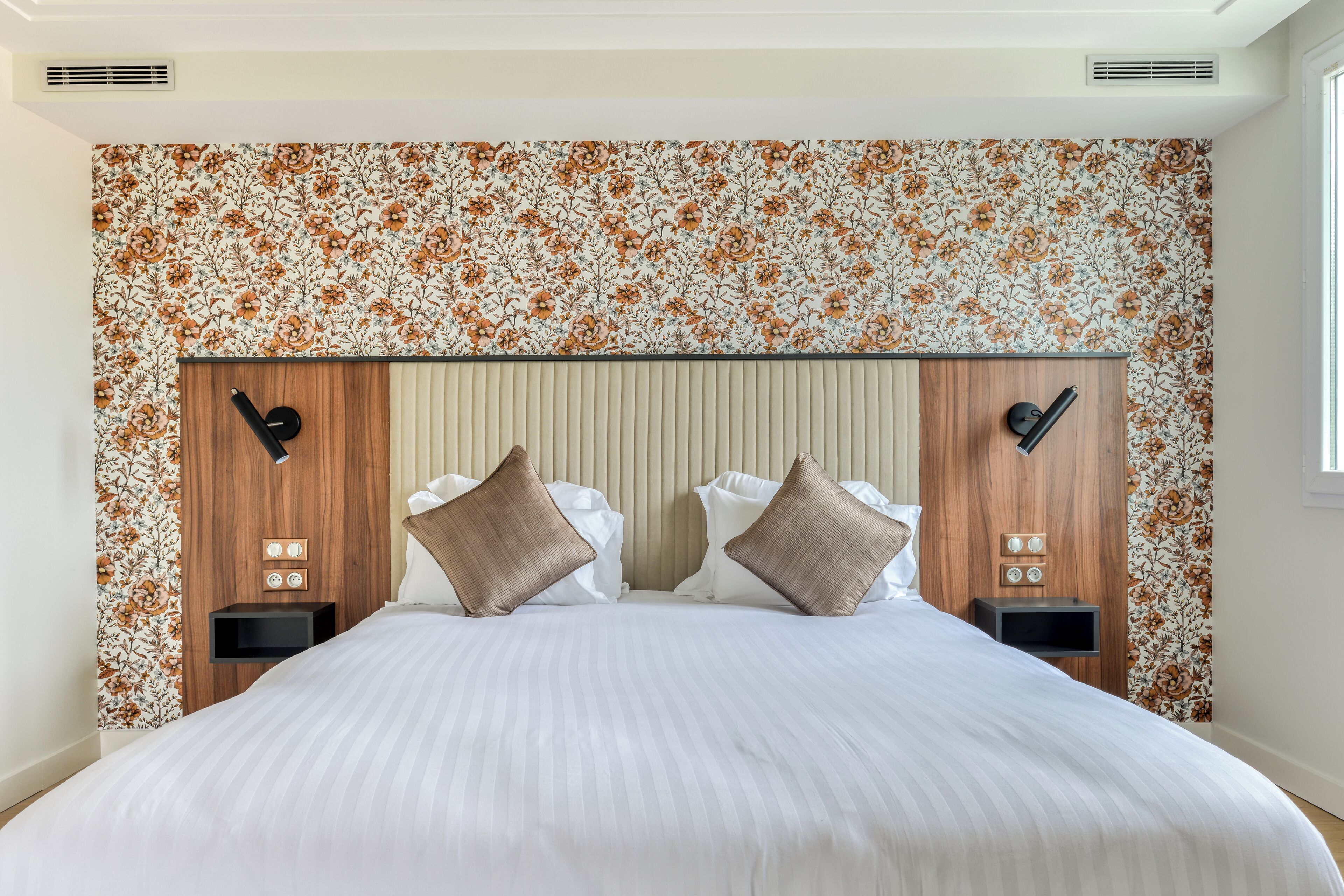 Una cama bien hecha con un edredón blanco a rayas, dos mesitas de noche de madera con relojes negros y un papel tapiz estampado con motivos florales.