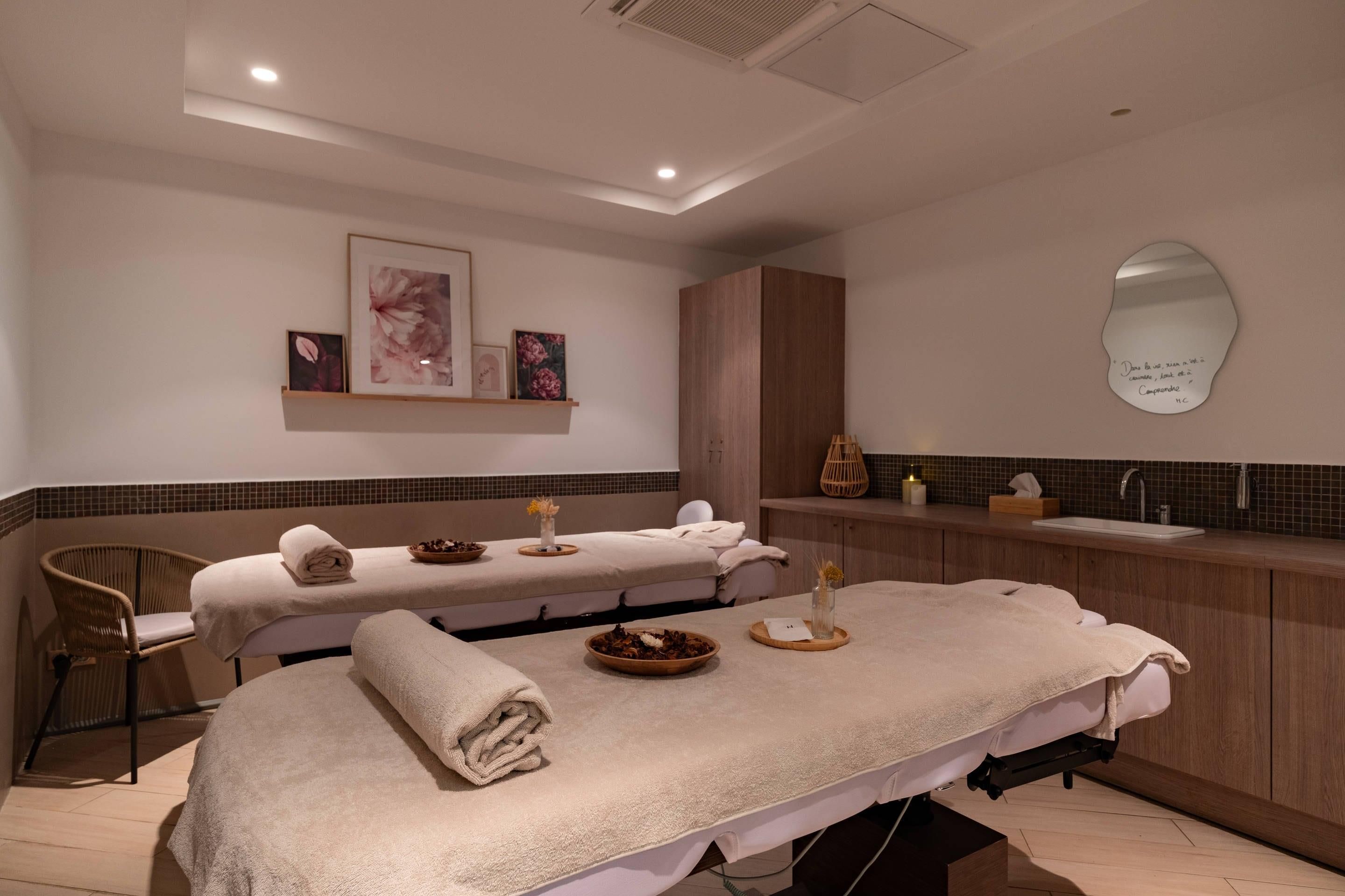 A spa room with two massage tables, a wooden cabinet, a mirror, and a wall-mounted shelf with framed artwork.