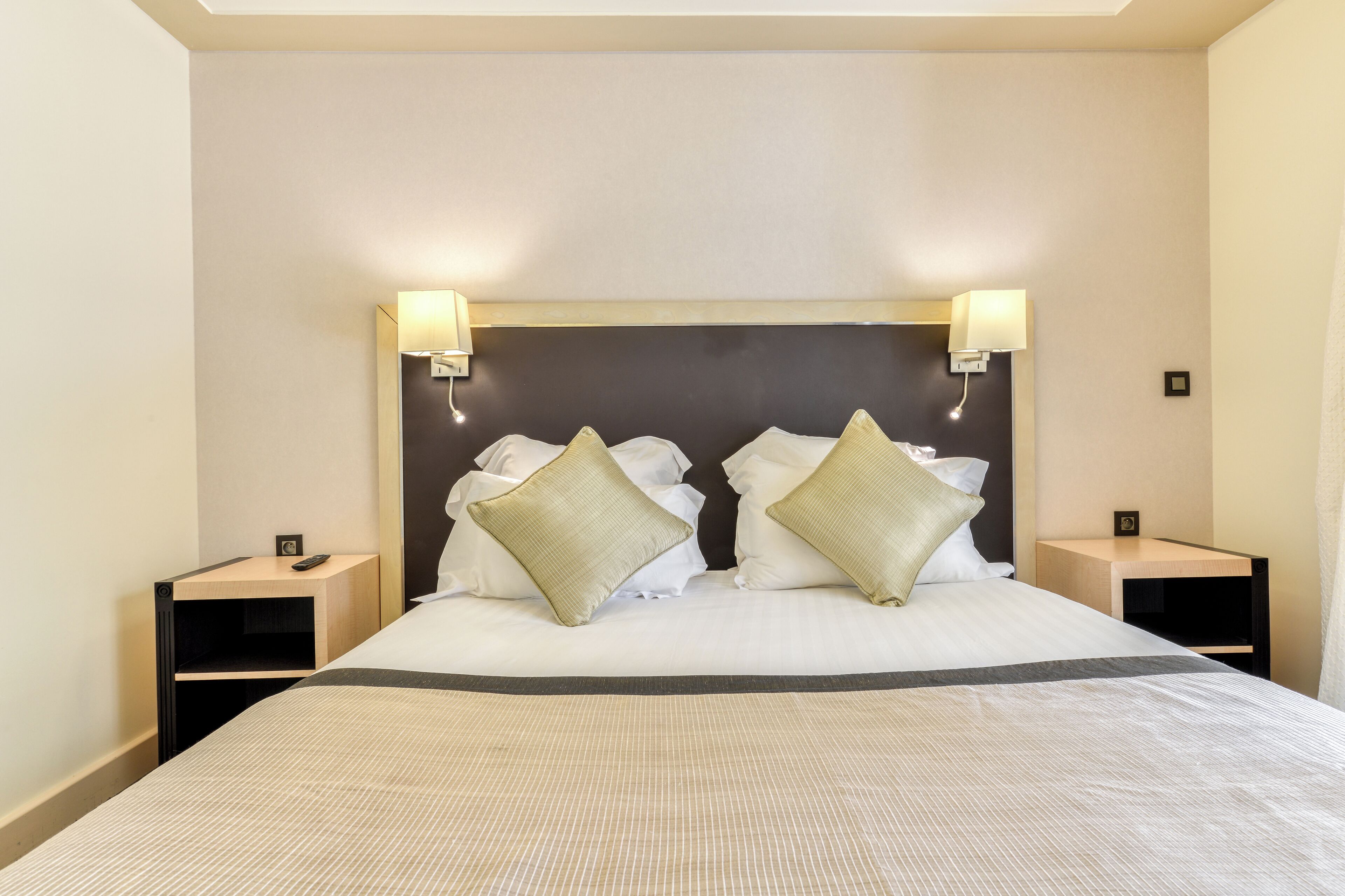 A neatly made bed with white linens and two pillows, flanked by bedside tables with lamps, in a room with plain walls and a simple headboard.