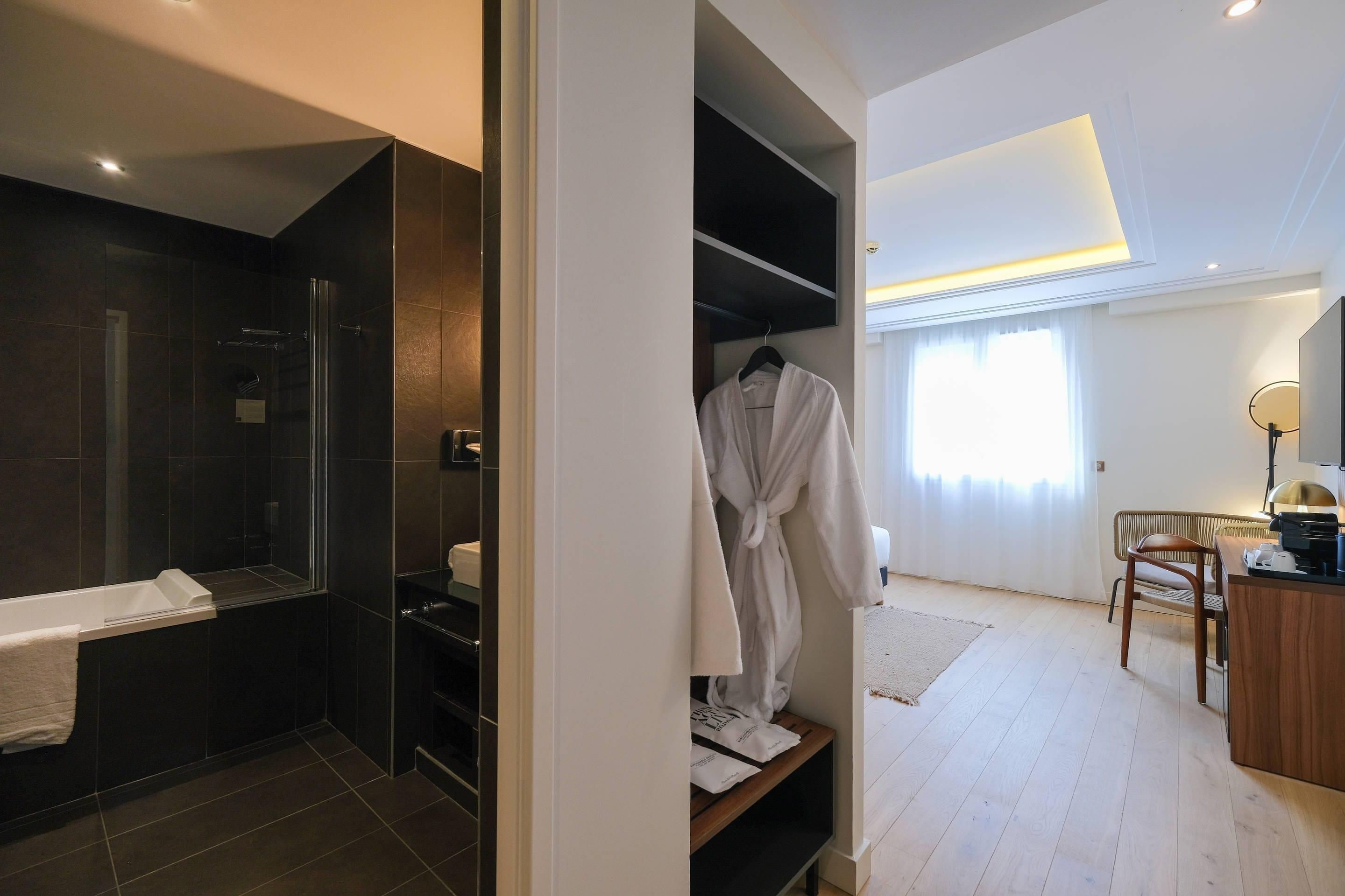 A modern bathroom with a glass shower enclosure, a white bathtub, and a walk-in closet with a robe hanging.