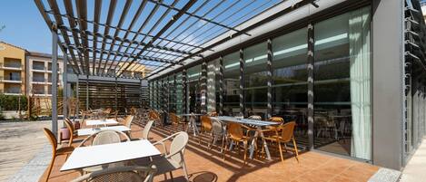 An outdoor patio with tables and chairs, a pergola, and large windows.