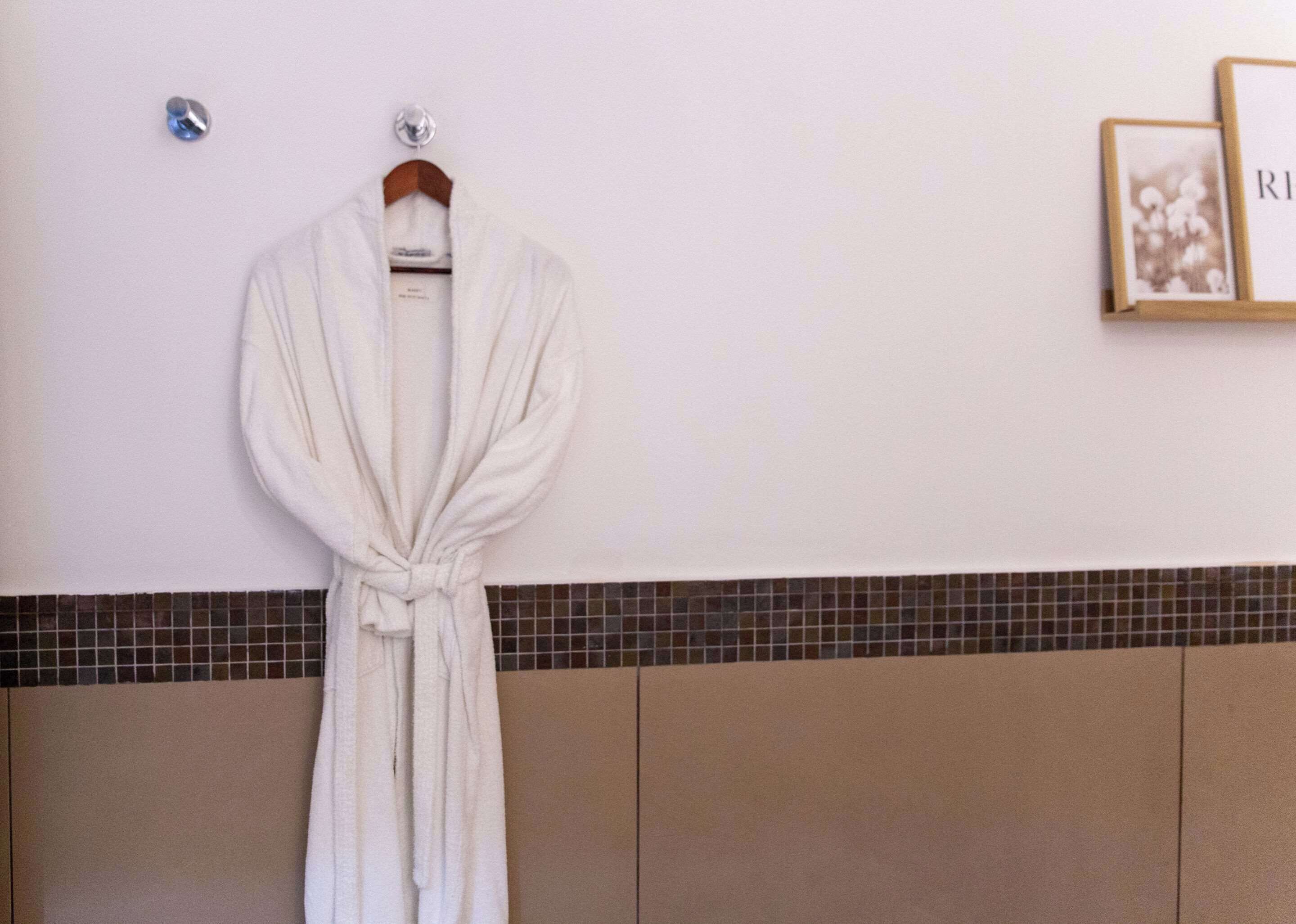 Un accappatoio bianco appeso a un appendiabiti in legno, su uno sfondo con una parete rivestita di piastrelle e un quadro appeso alla destra.