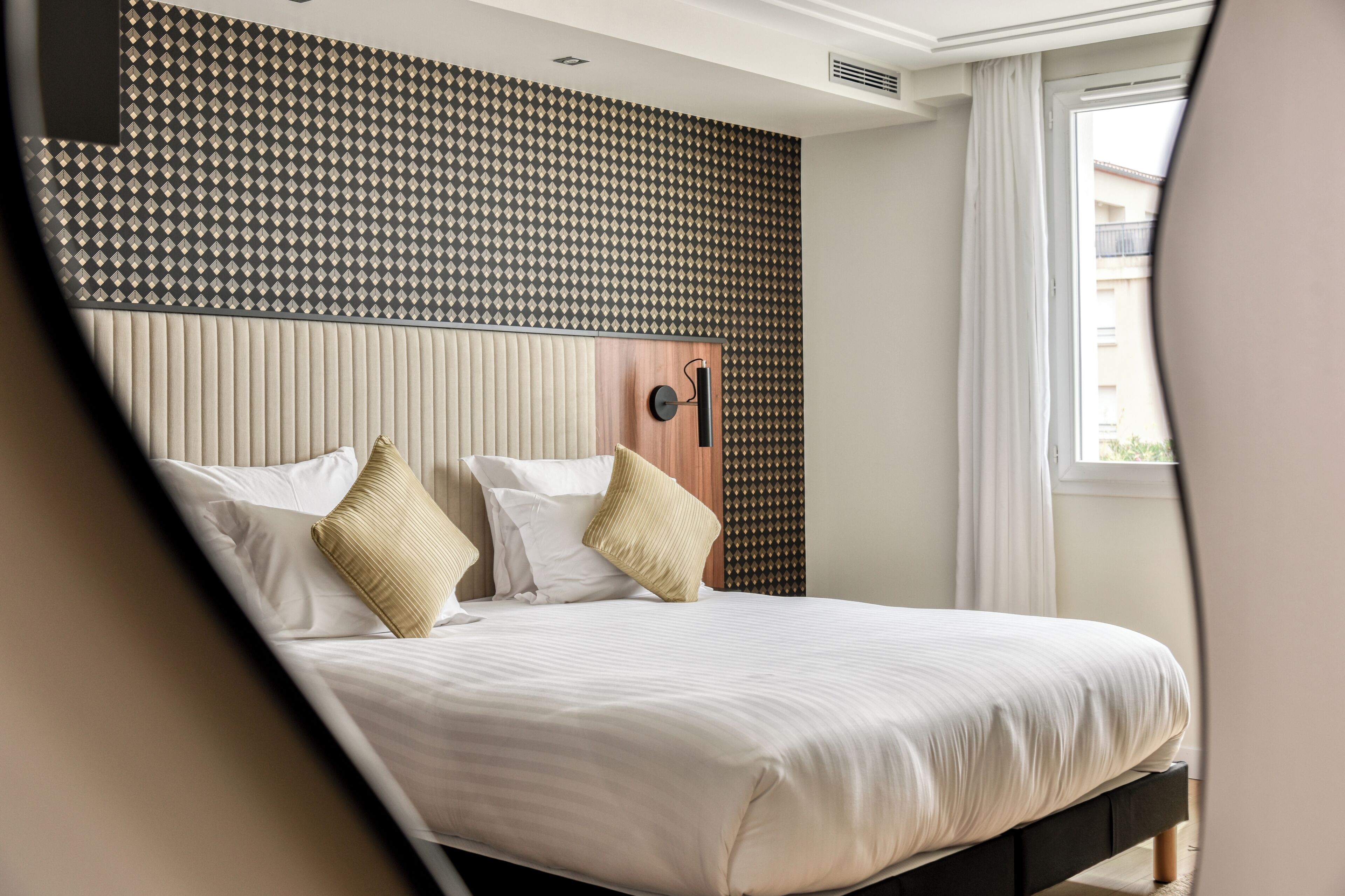 Een net opgemaakt bed met witte lakens en beige kussens in een hotelkamer, met een behang en een raam met gordijnen.