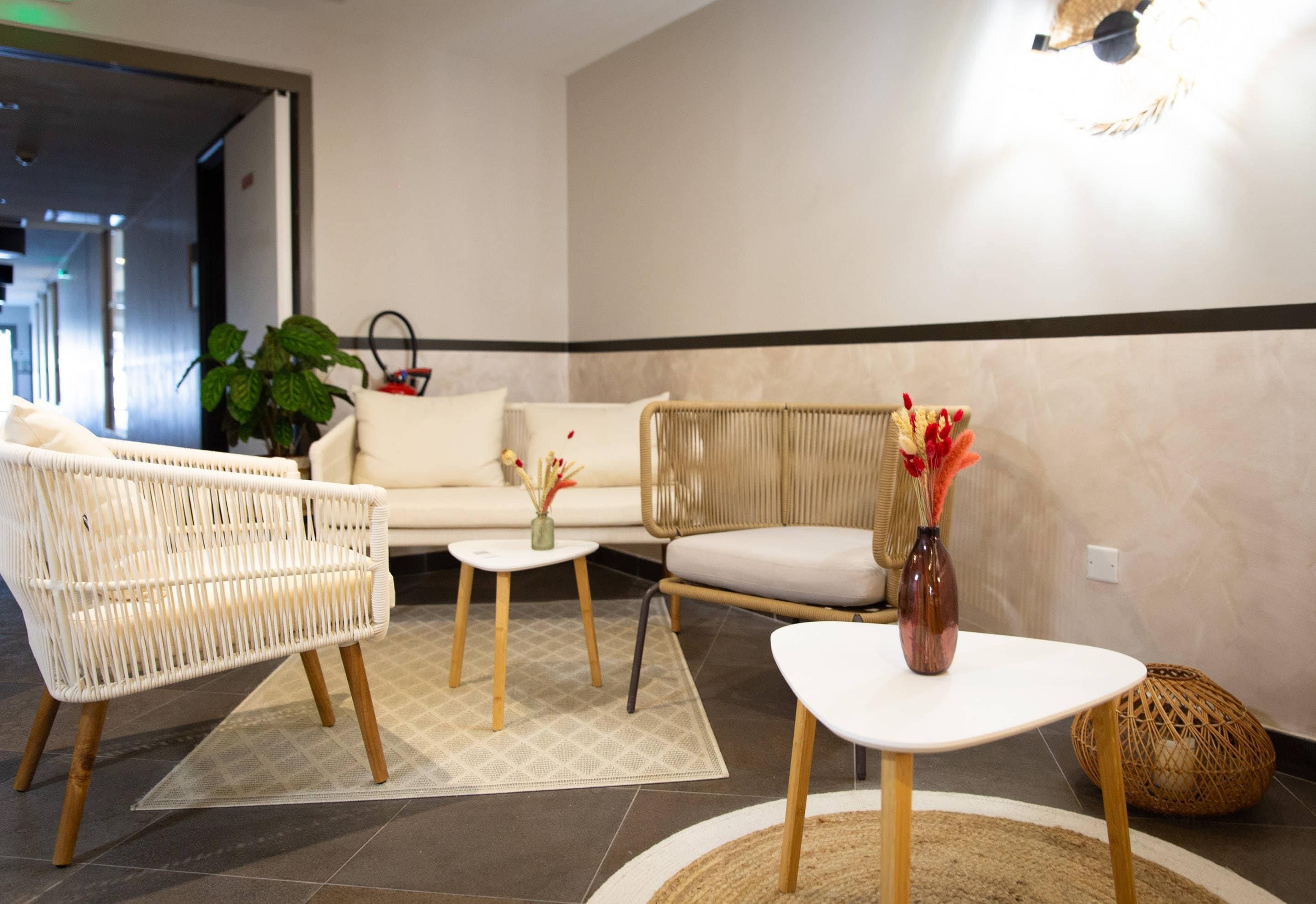 Un hall d'hôtel moderne avec un canapé blanc, des fauteuils en osier, une petite table surmontée d'un vase et un appliques murales décoratives.