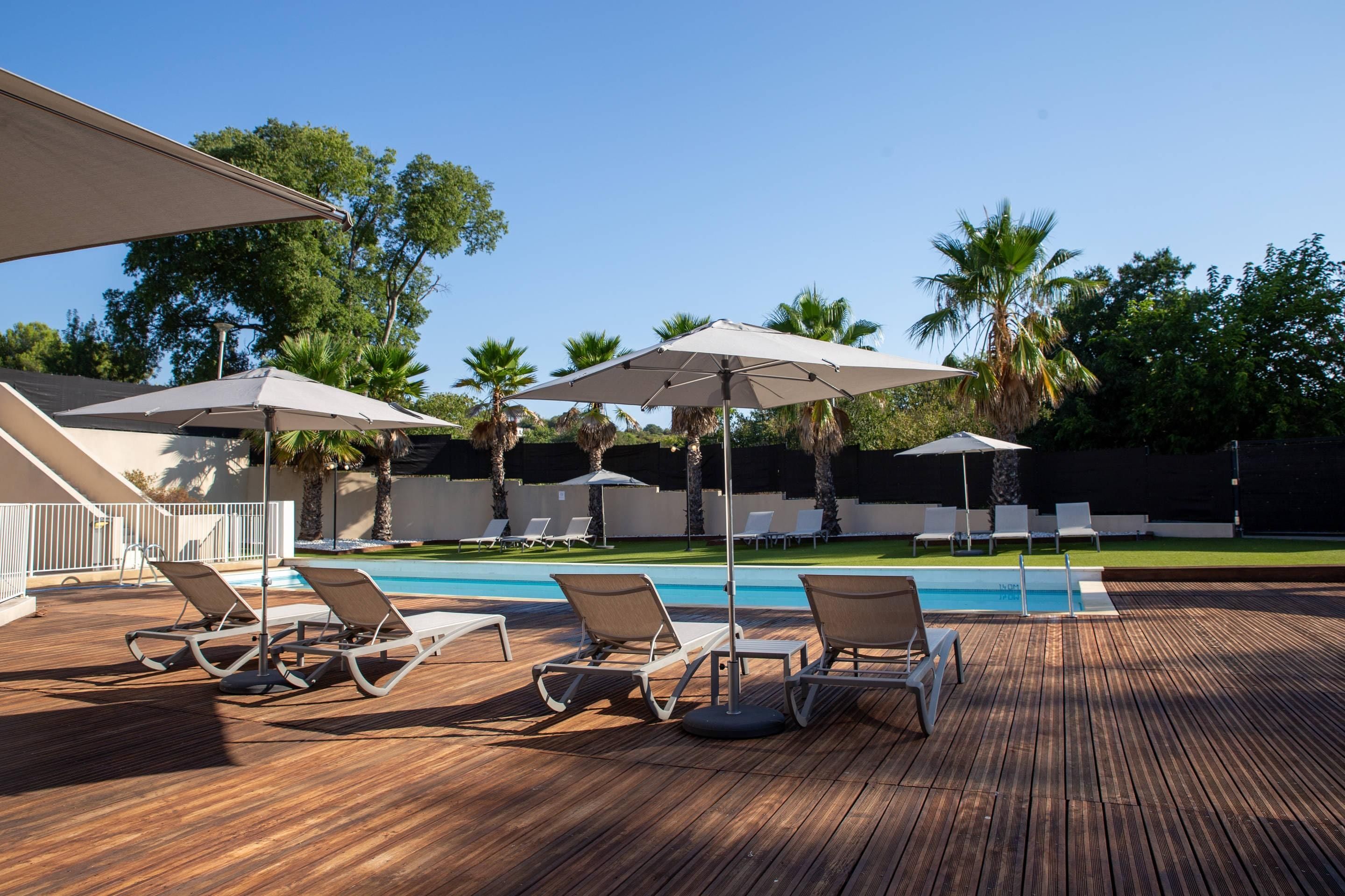 A poolside area with lounge chairs and umbrellas, surrounded by palm trees and a clear sky.