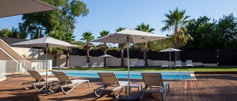 A poolside area with lounge chairs and umbrellas, surrounded by palm trees and a clear sky.