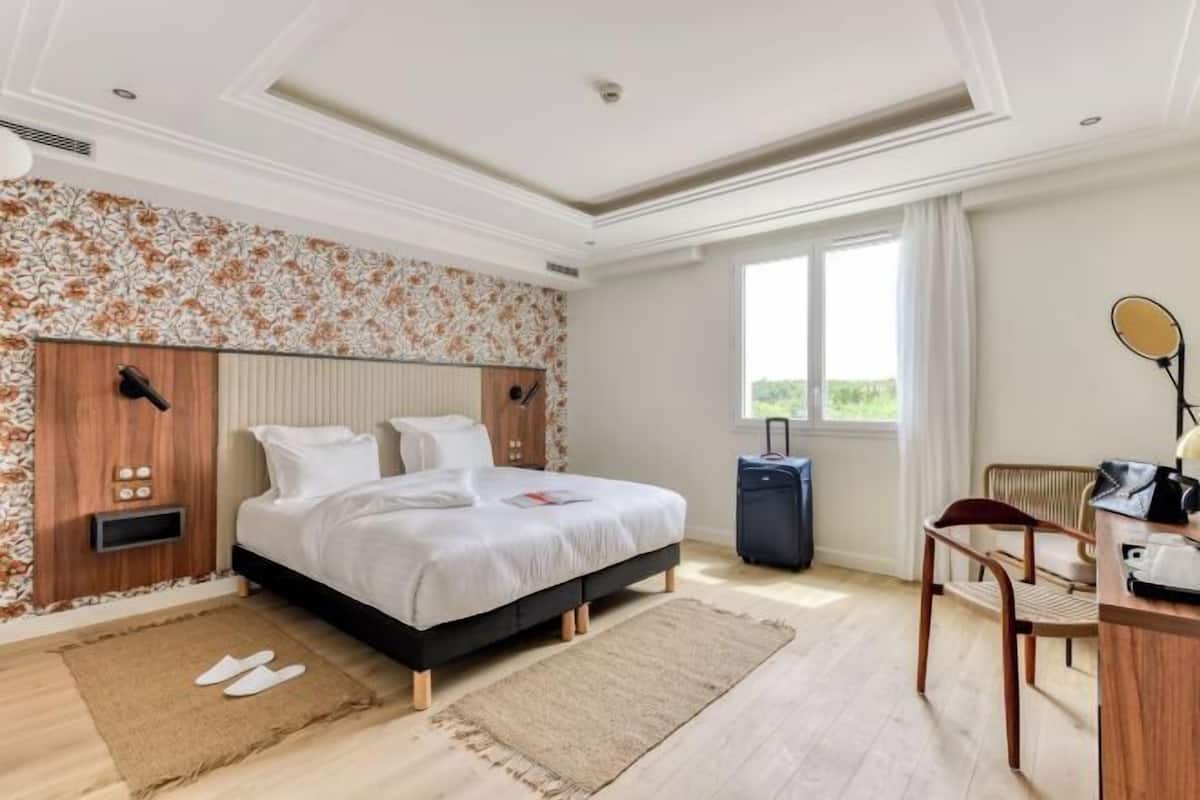 A hotel room with a bed, a desk, a chair, a suitcase, and a window with a view.