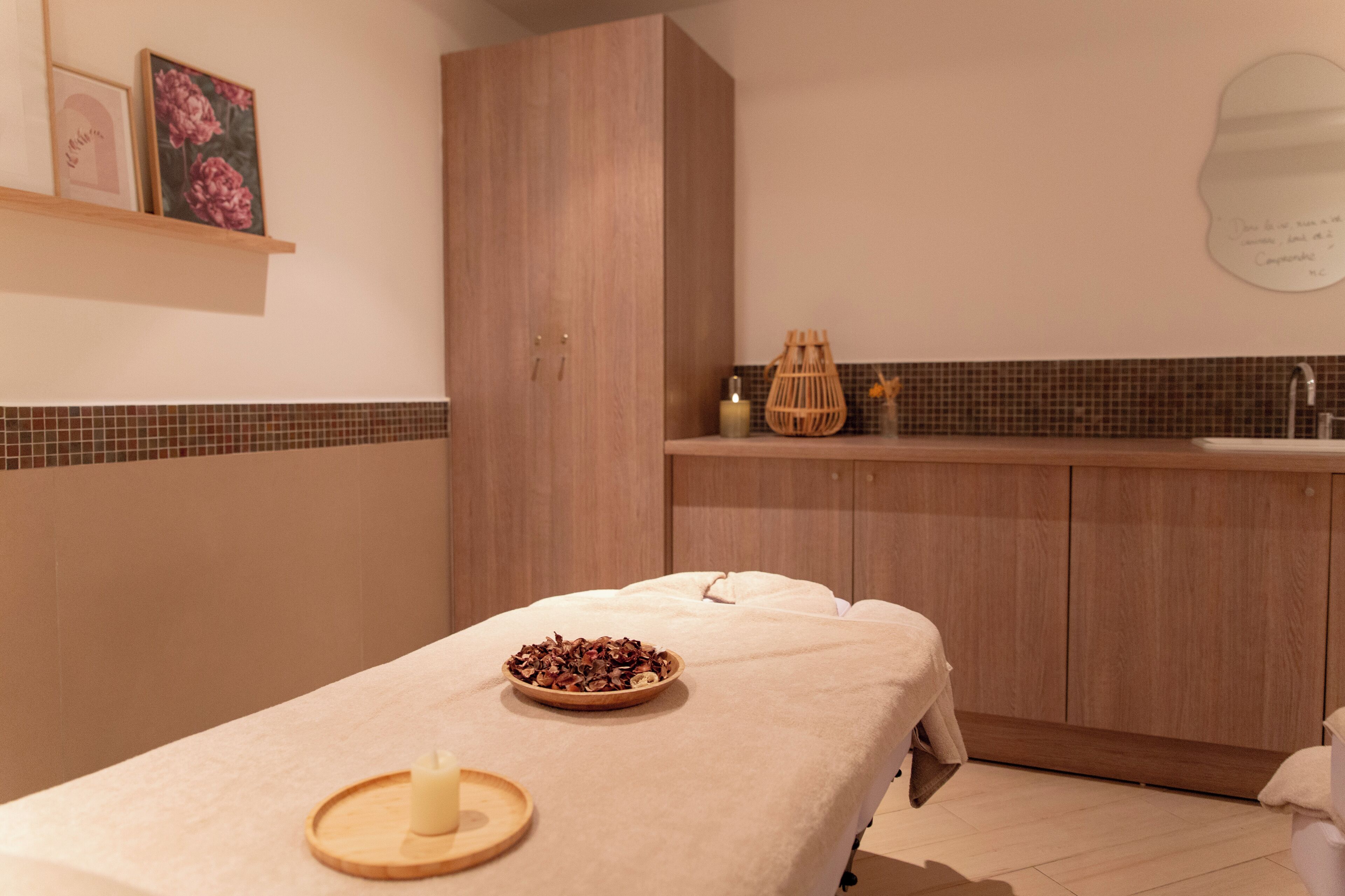 A massage table with candles and a bowl of dried flowers, wooden cabinets, a tiled backsplash, and a small shelf with decorative items.